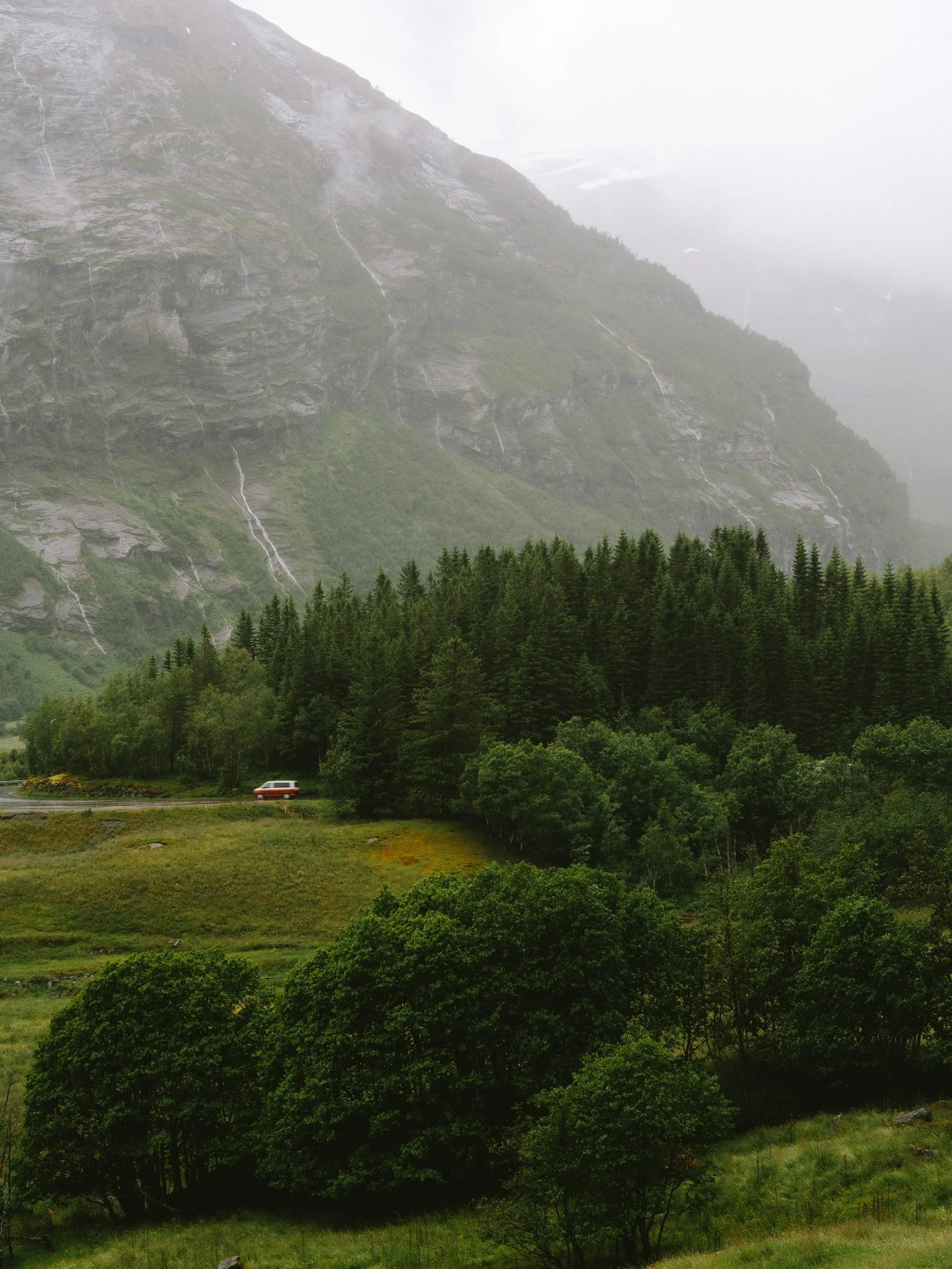 A landscape with a mountain, lush green trees, and a small vehicle on a winding road.