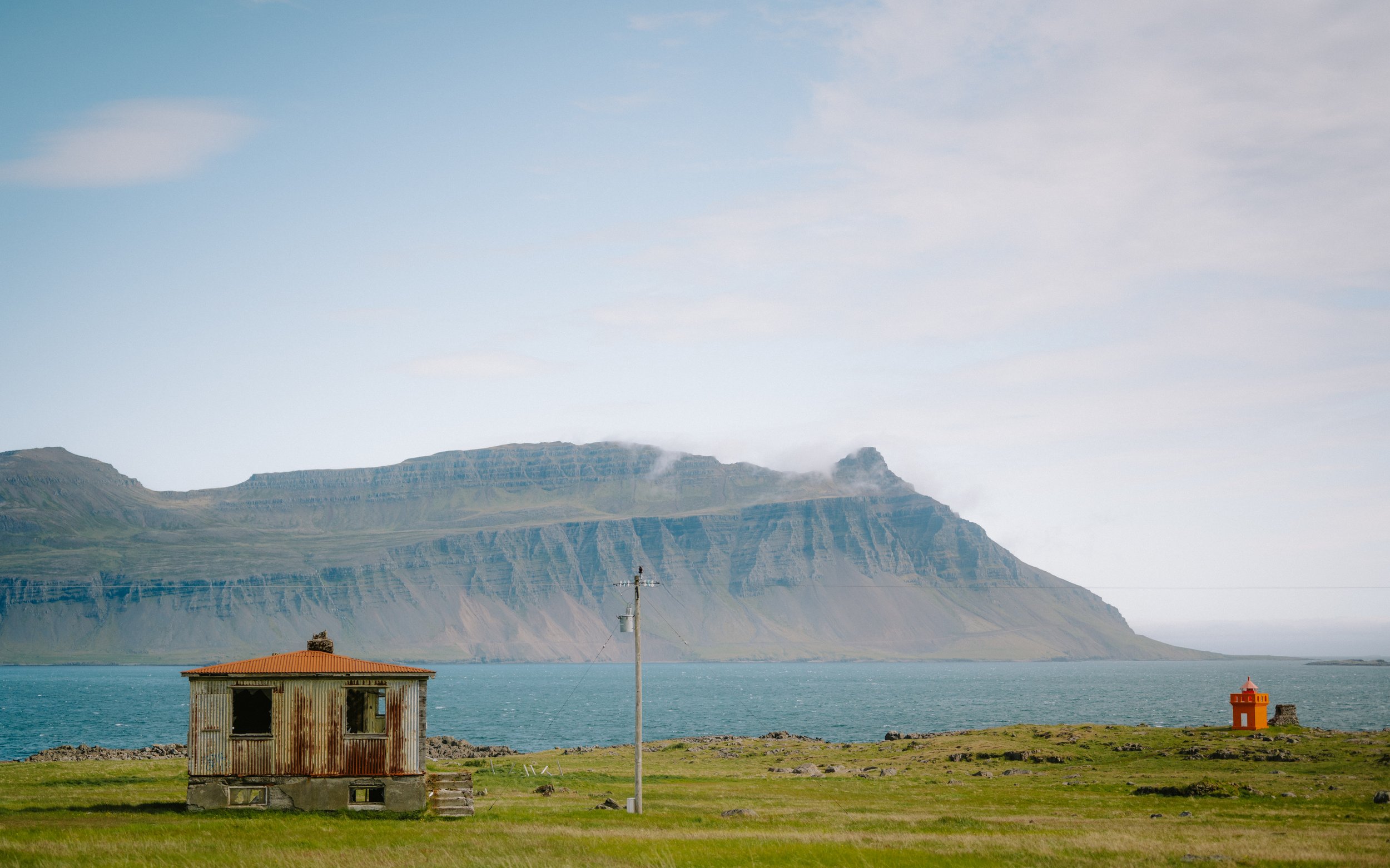 A landscape featuring a grassy field with two small structures: one rusty, abandoned house and a red lighthouse near a body of water, with mountains in the background and a partly cloudy sky.