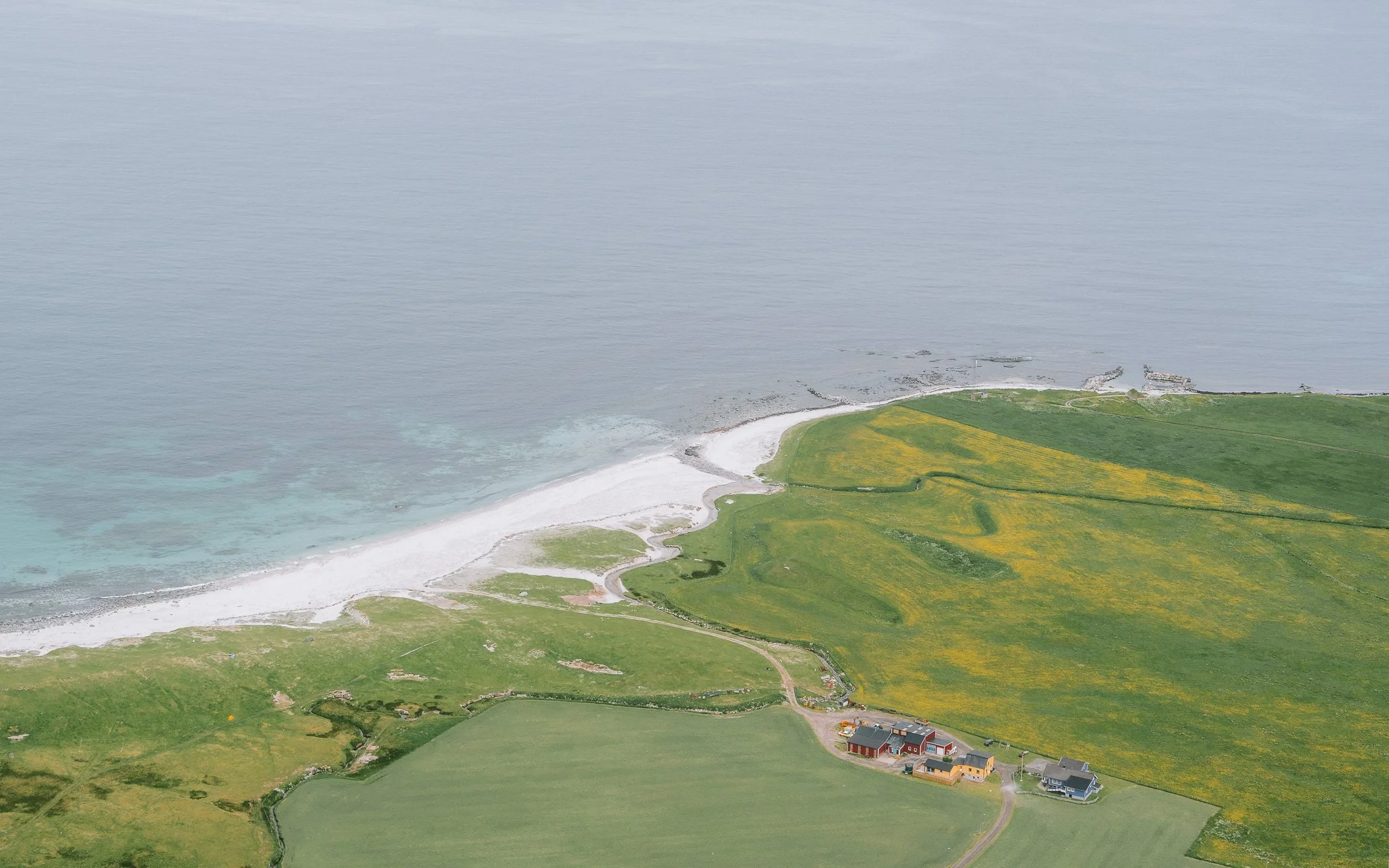 Aerial view of a coastal landscape featuring a white sandy beach, rocky shoreline, and green fields with a few scattered houses near the shore.