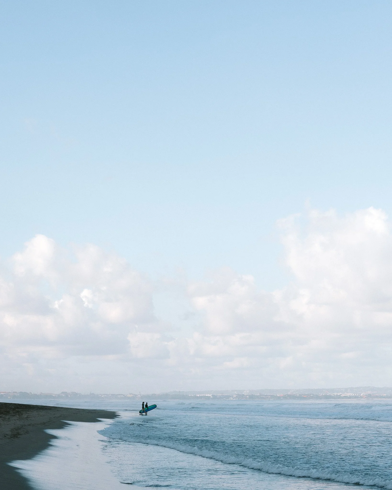Two people walking on the beach with a surfboard near the shoreline, under a cloudy sky.