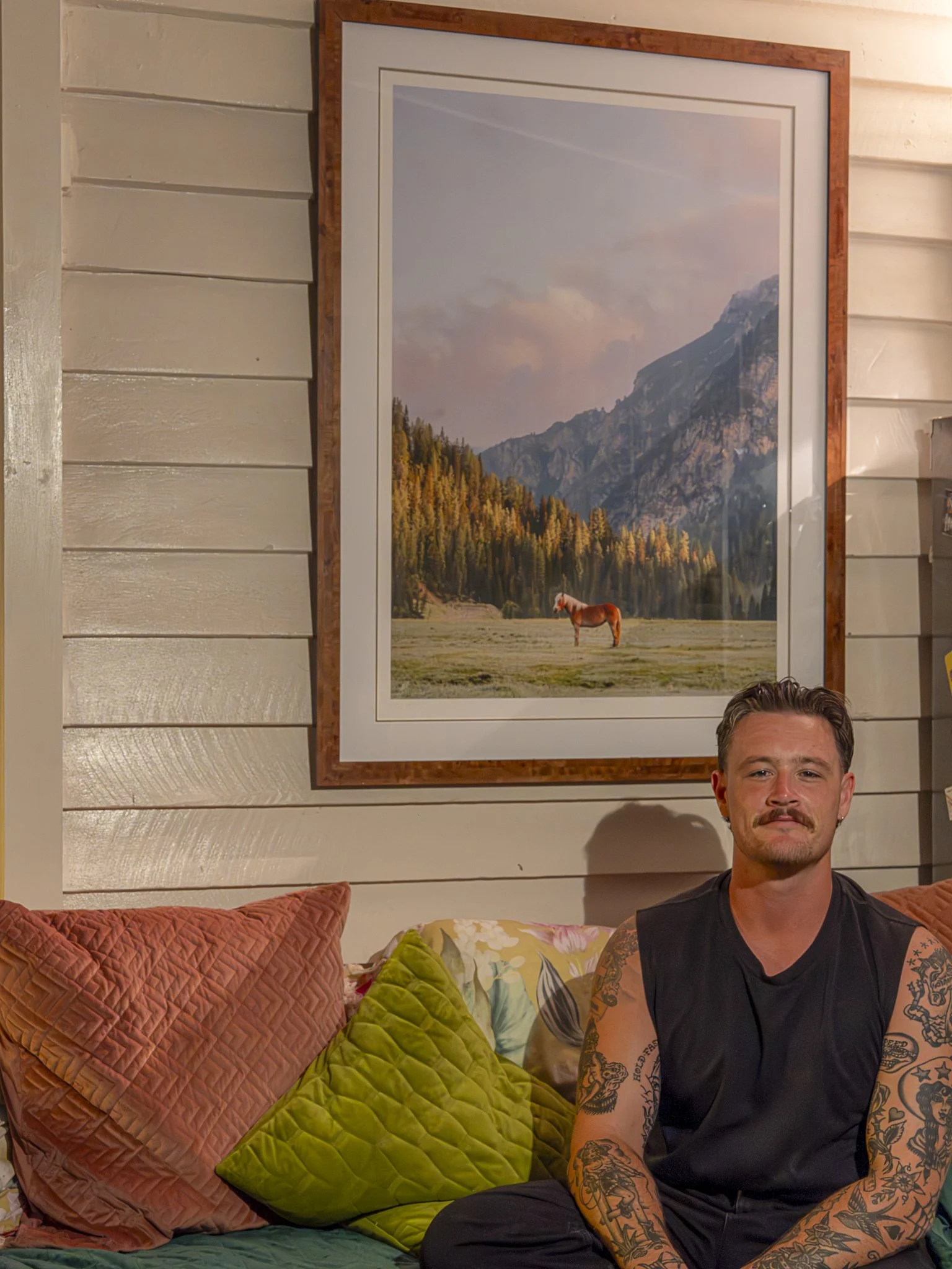 A man with tattoos on his arms sitting on a couch with colorful pillows, in front of a wall with horizontal wooden planks and a large framed landscape photograph of mountains, trees, and a horse.