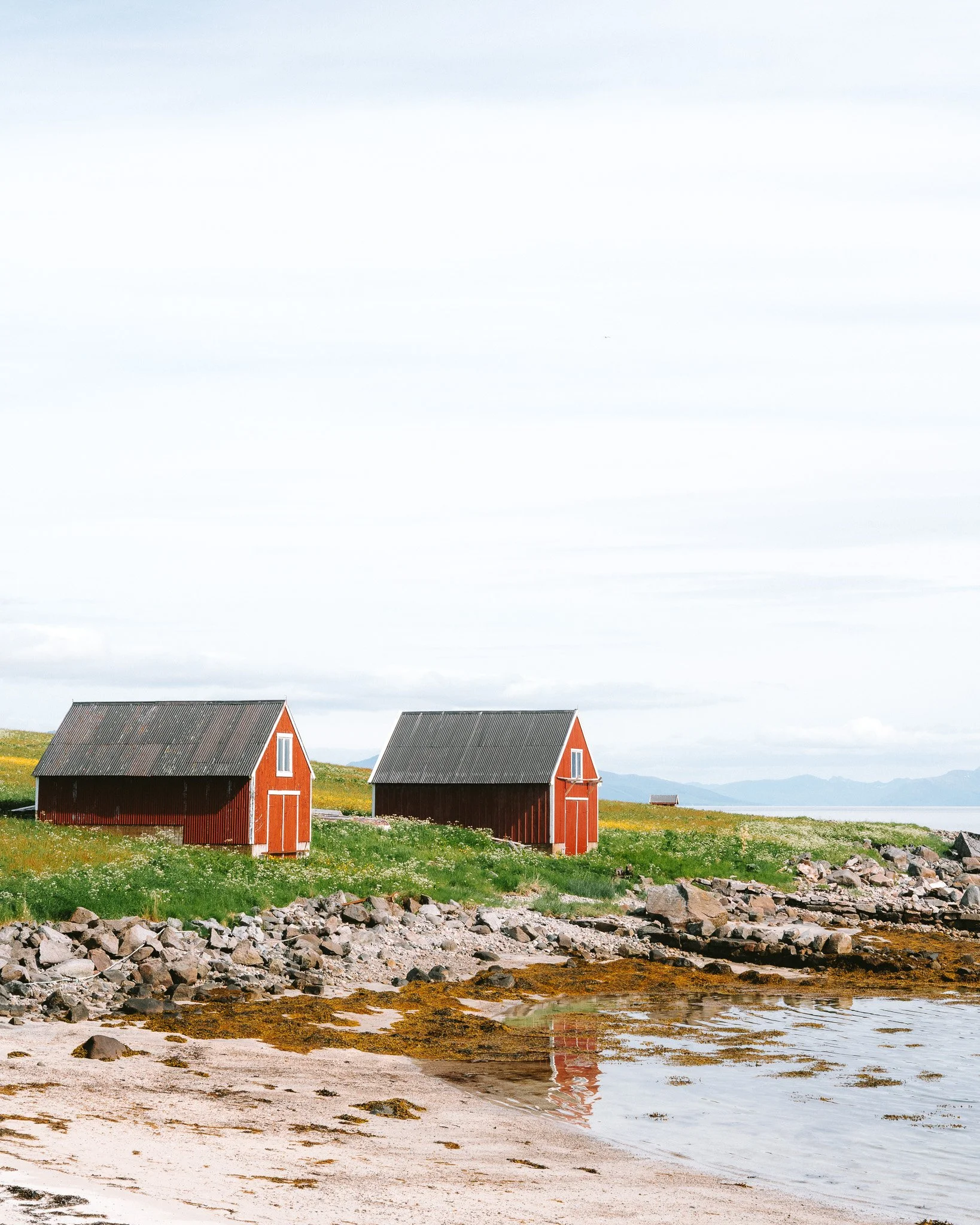 Two red beach cabins on a grassy shoreline with rocks and water in the foreground, overcast sky in the background.