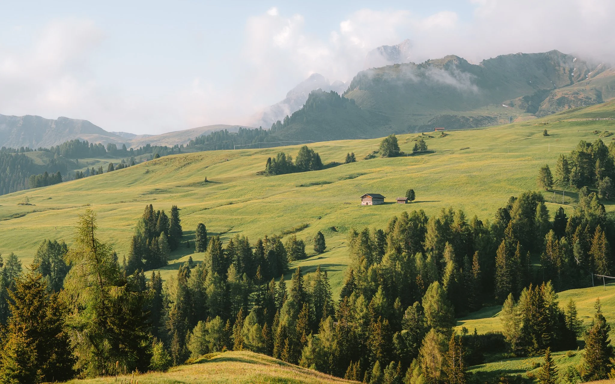 Green rolling hills with scattered trees and small houses, mountains in the background under partly cloudy sky.