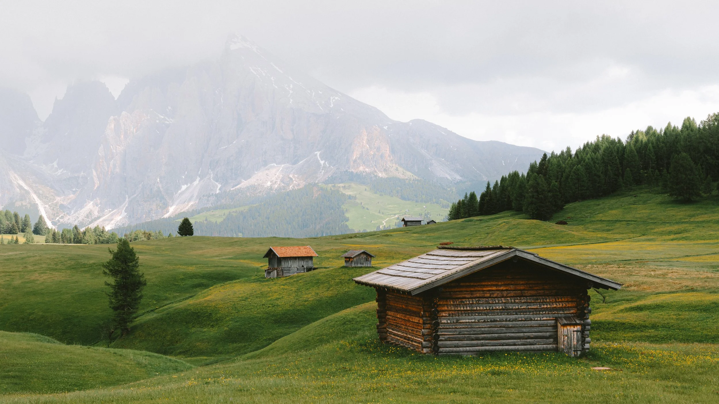 A pastoral landscape with rolling green hills, a rustic wooden cabin in the foreground, and several small wooden houses scattered across the hills. In the background, there are tall trees and towering snow-capped mountains under a cloudy sky.
