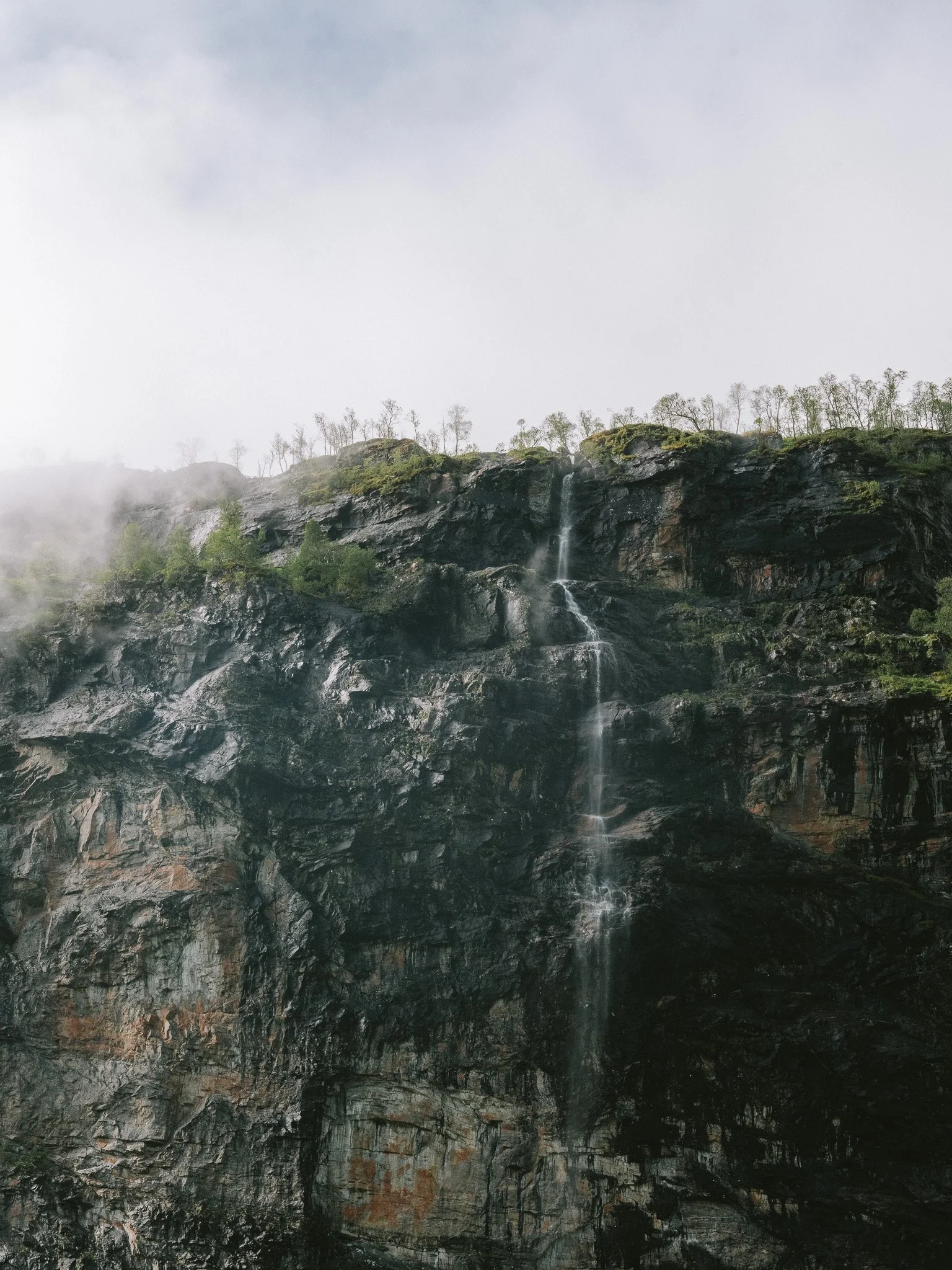 Waterfall cascading down a dark rocky cliffside with trees at the top, mist and clouds in the sky.