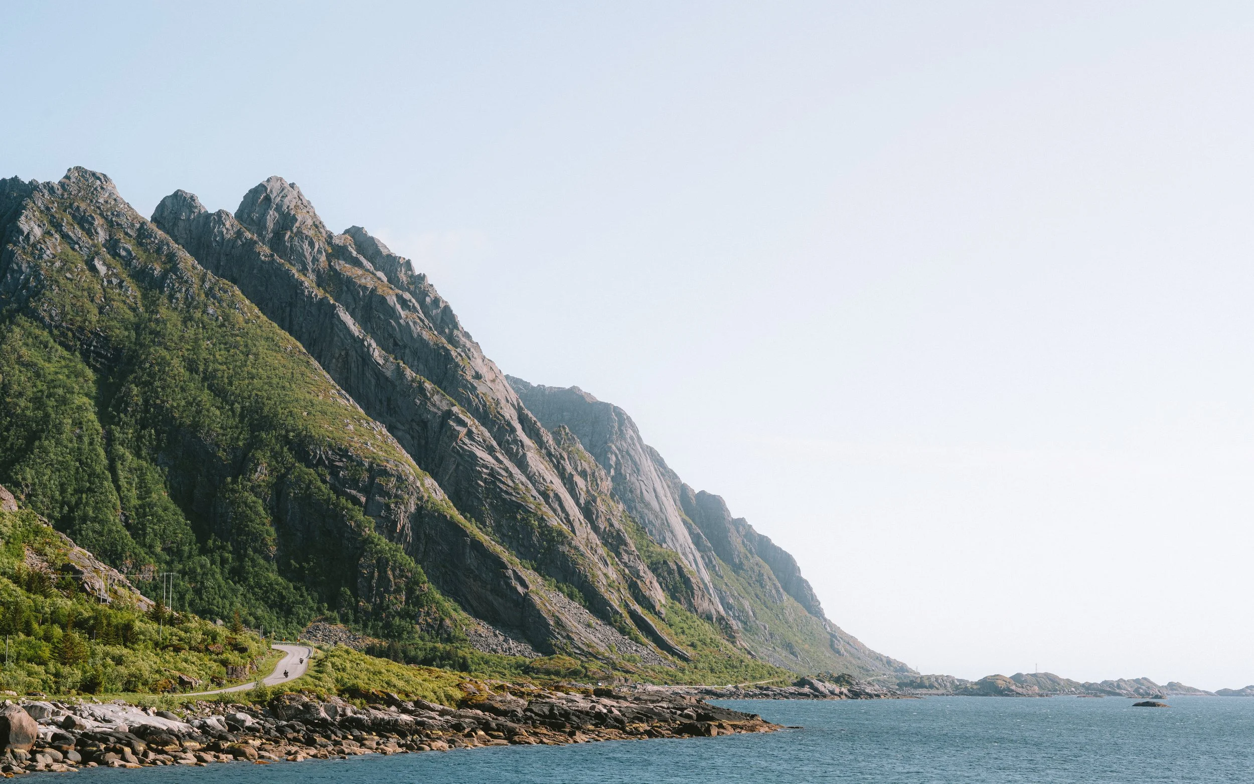 Coastal landscape with towering green mountains and a rocky shoreline beside the ocean under a clear sky.