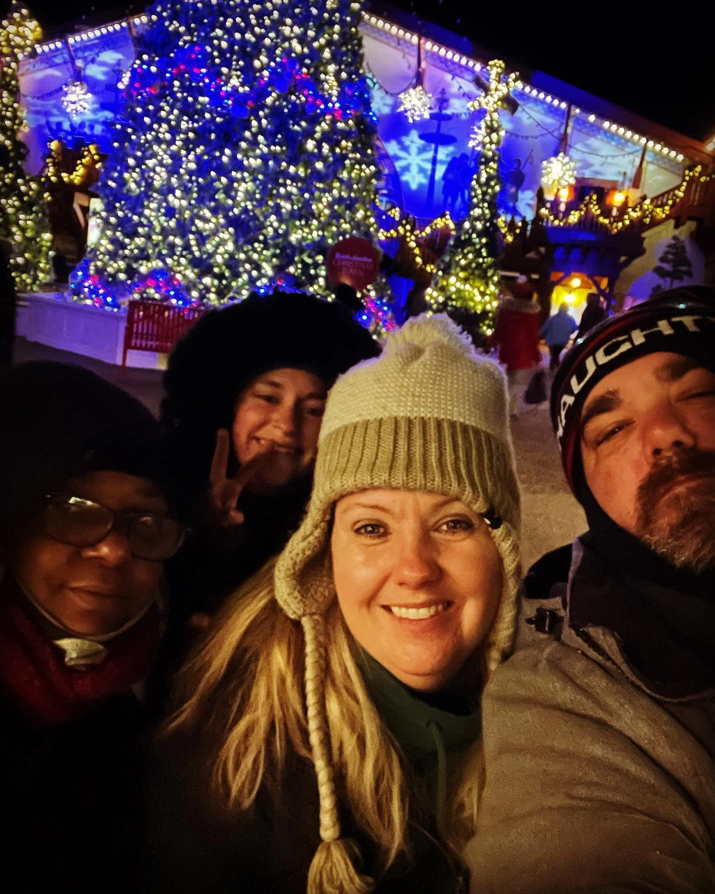Group of four people taking a selfie in front of a decorated Christmas tree at night, with holiday lights and snowflake decorations in the background.