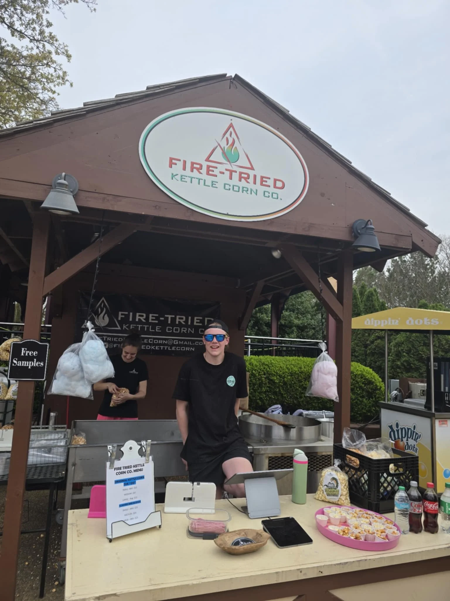 A food stall named 'Fire-Tried Kettle Corn Co.' at an outdoor event, with two smiling people behind the counter. The stall offers kettle corn and has a sign for free samples. There is popcorn in bags hanging and on the table, with drinks and small sn