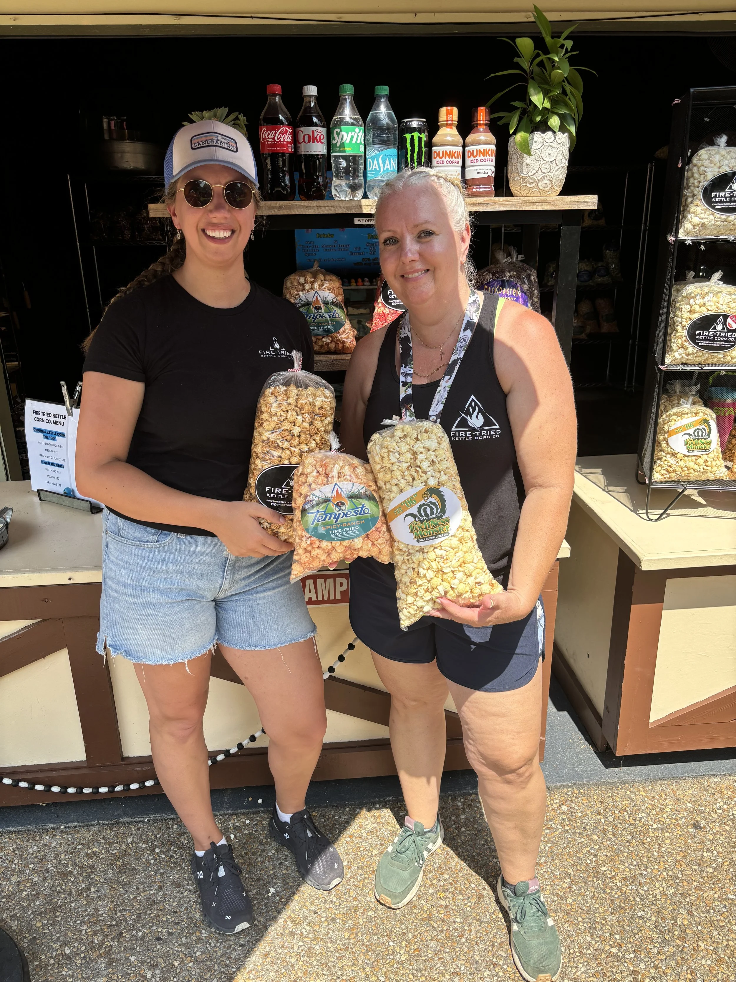 Two women standing in front of a snack stand holding large bags of caramel popcorn, with soda bottles and a potted plant behind them.