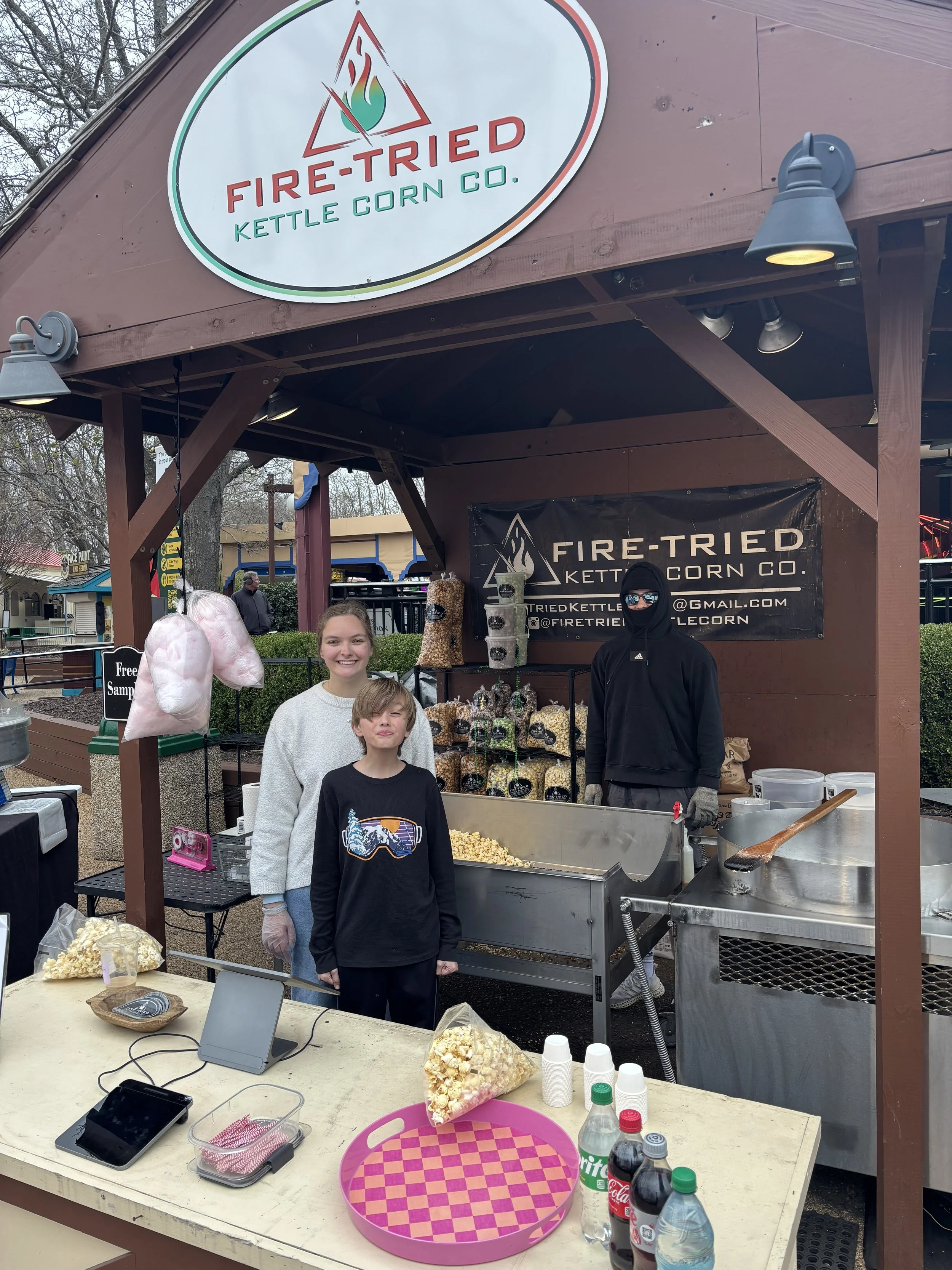 A food stand named Fire-Tried Kettle Corn Co. with two children and a vendor making kettle corn. The stand has a large sign with a flame logo. The children are smiling, and the vendor wears sunglasses, a mask, and gloves while preparing popcorn. Tabl
