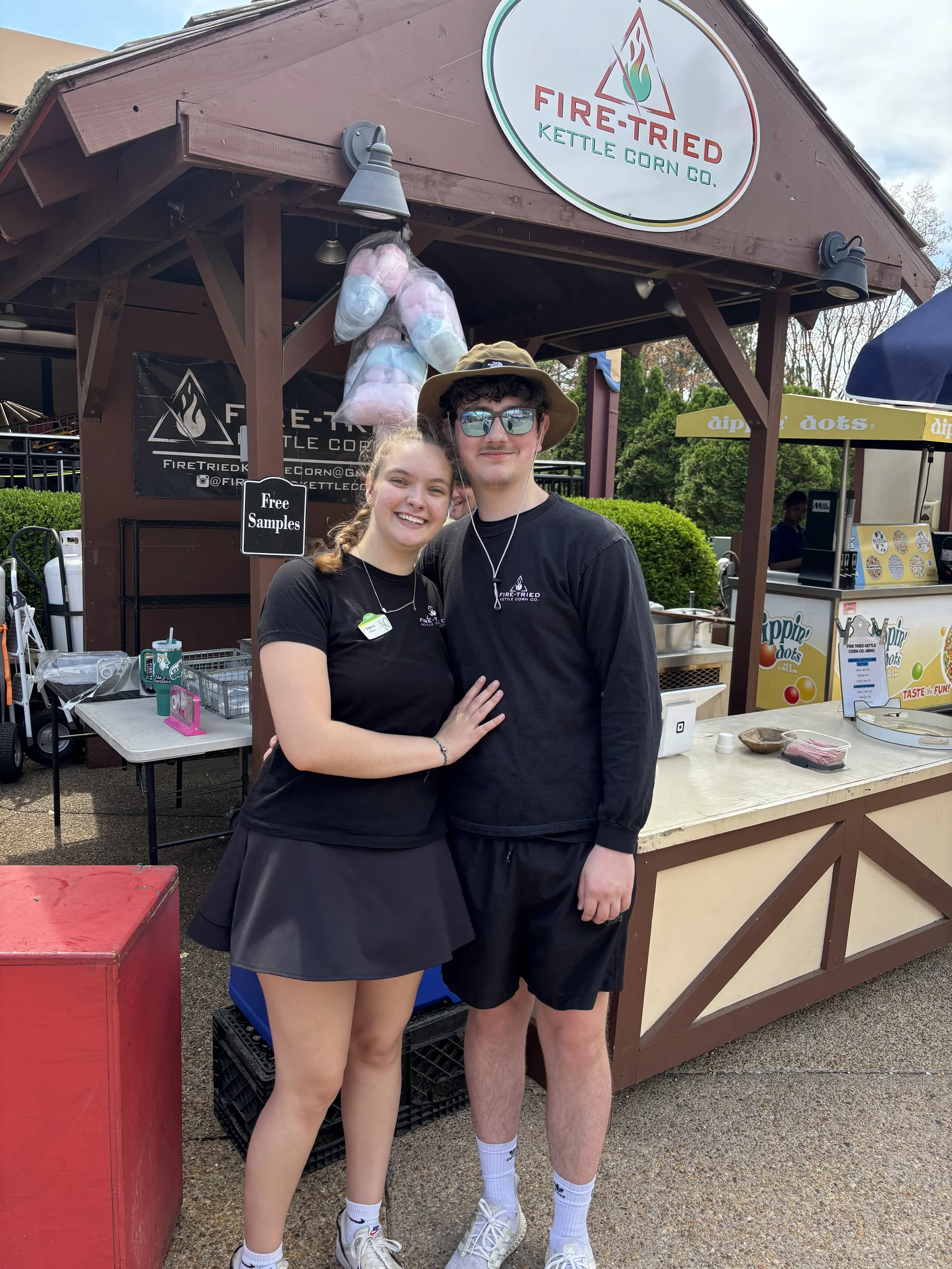 A young woman and a young man smiling and posing together at a fair food stand, with the young woman touching the young man's chest. The stand has a sign reading 'Fire-Trusted Kettle Corn Co.' and cotton candy hanging from the top.