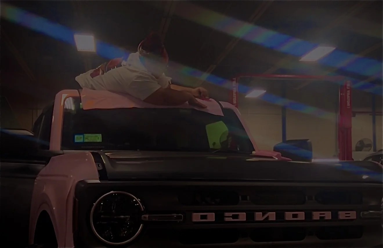 A person working on a pink and black sports car inside a garage or workshop, with cars on lifts visible in the background.