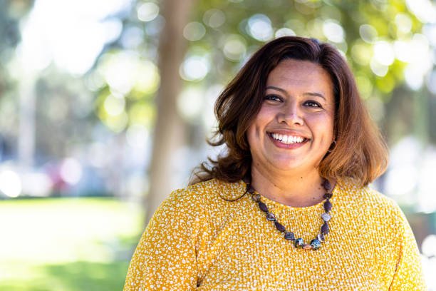 Woman smiling outdoors wearing a yellow patterned top and a beaded necklace.