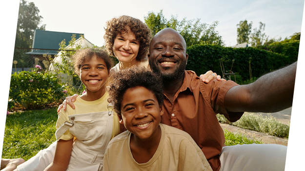 A family of four taking a selfie outdoors in a garden with green trees and a house in the background. They are smiling and appear happy.