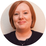 Close-up of a woman with short red hair, smiling, wearing a black top with a necklace, against a neutral background.