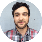 Portrait of a young man with dark hair and a beard, wearing a plaid shirt, smiling with a light blue background.