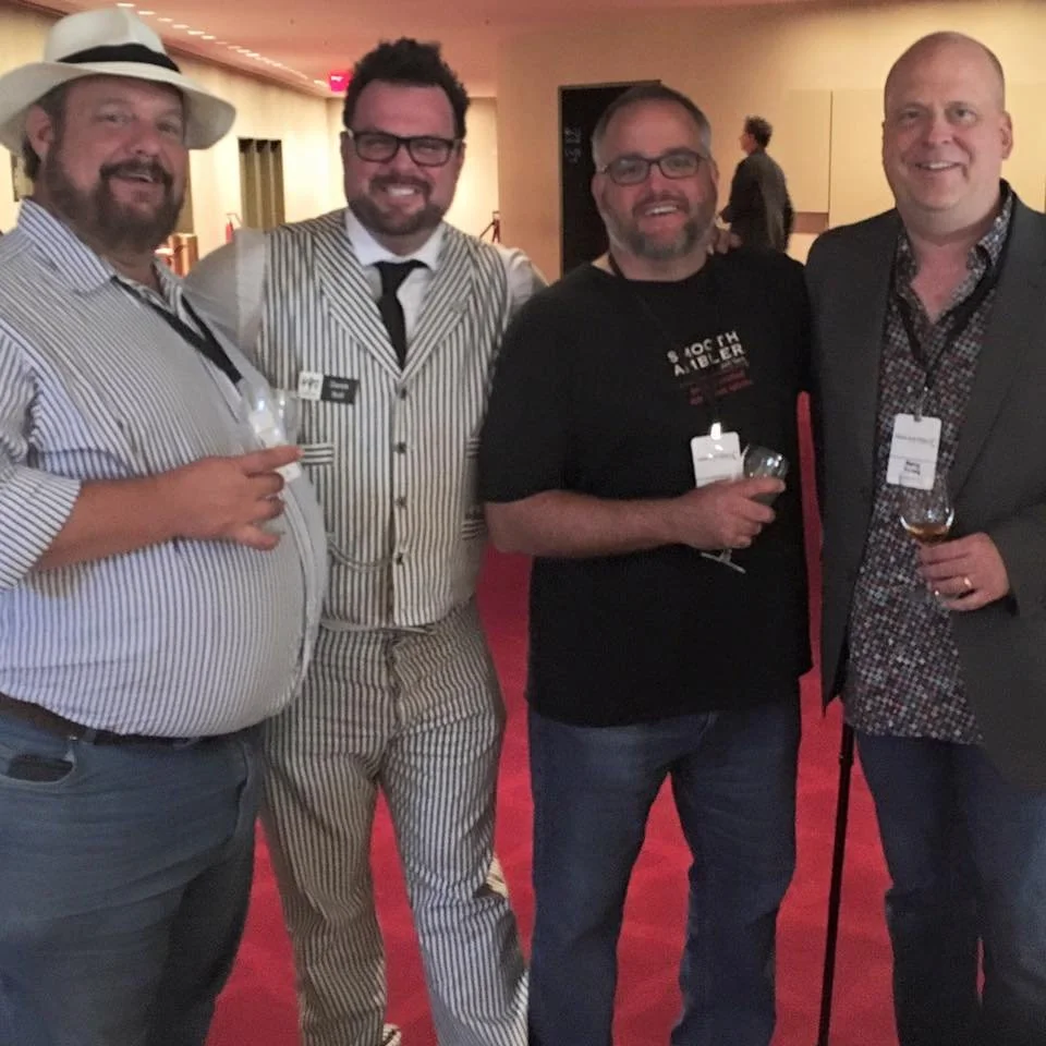 Four men standing together at a social event, each holding a glass of wine or drink, smiling at the camera, in an indoor setting with a red carpet and warm lighting.