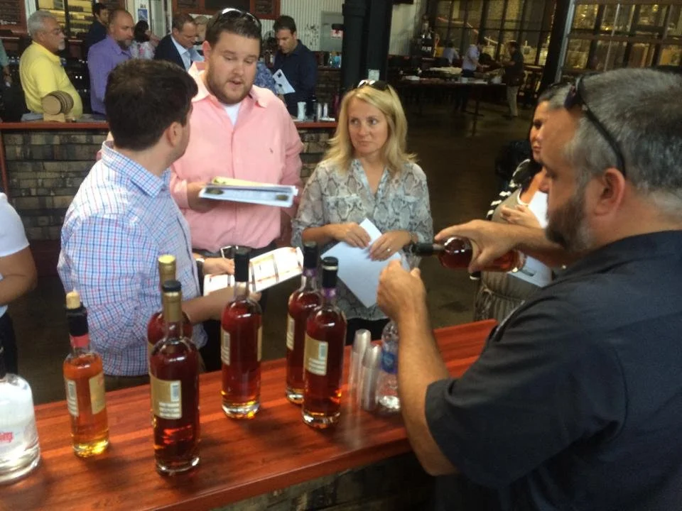 A man with a beard and glasses pouring a dark liquor into a glass at a tasting event with four bottles of liquor on a wooden table, while several people listen and hold notebooks inside a busy indoor venue.