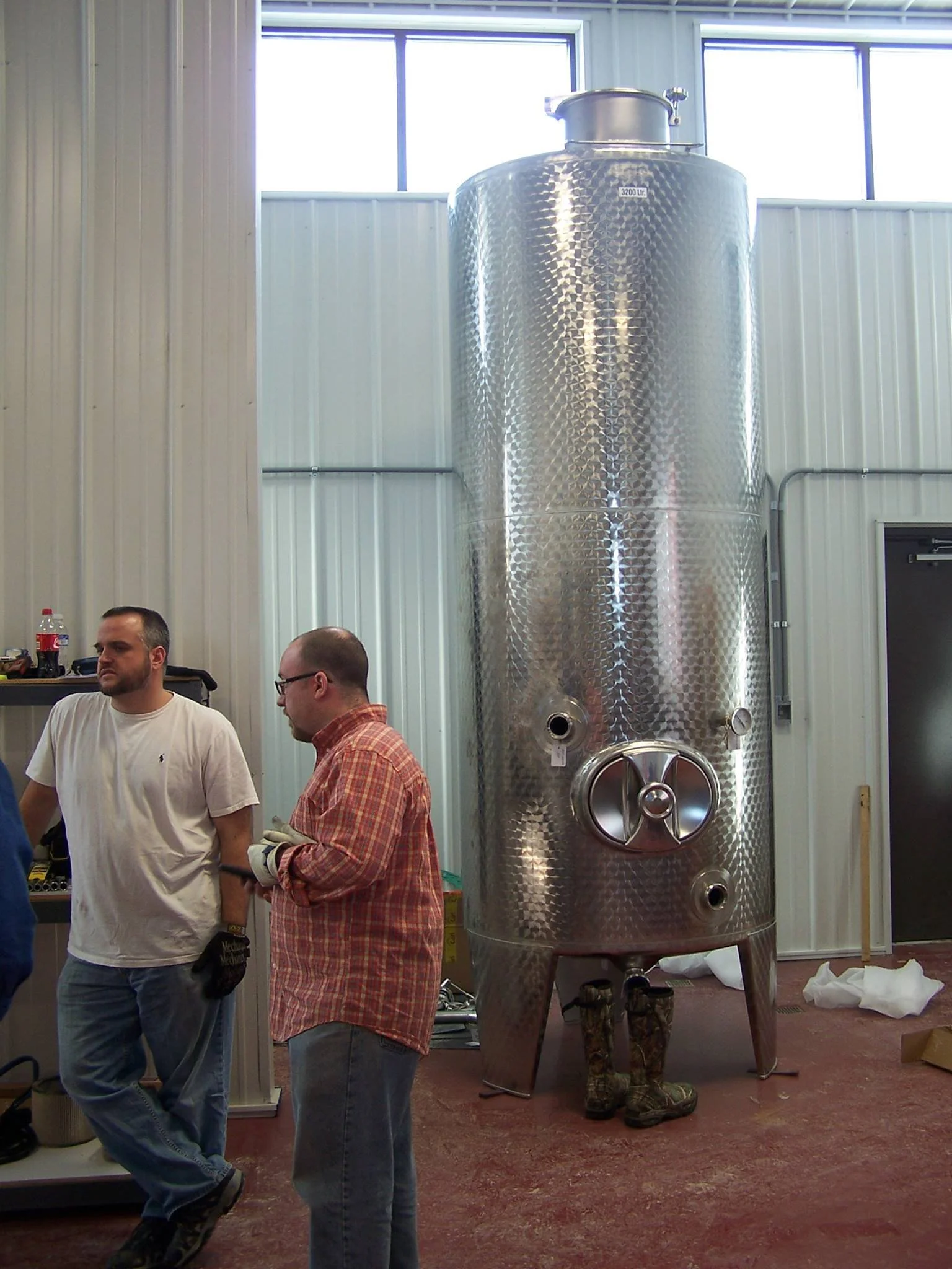 Two men standing and conversing next to a large stainless steel cylindrical fermentation tank in a brewery. The tank has a diamond-patterned surface and several valves, with a pair of boots visible underneath it. The brewery has white walls, large wi