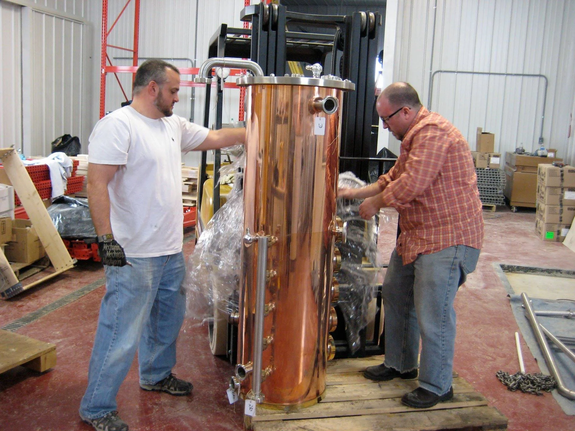 Two men working on a large copper distillation column in a warehouse or industrial setting.