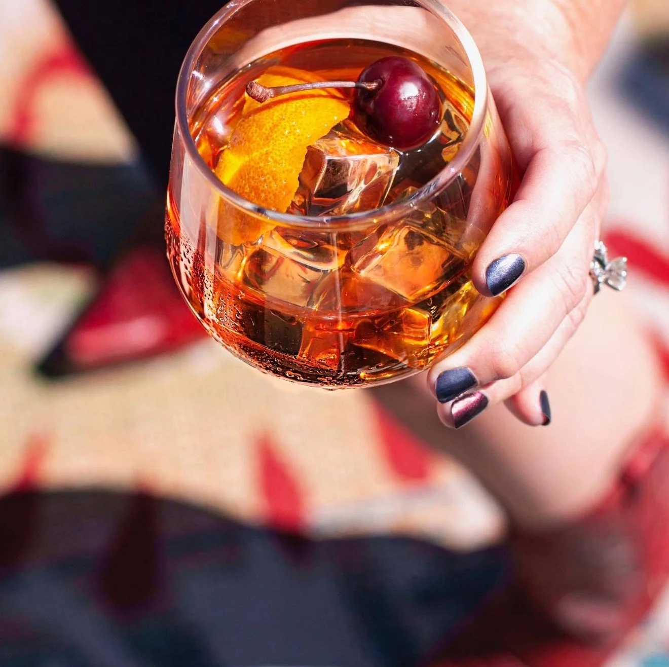 A hand holding a glass of amber-colored cocktail with ice, a cherry, and an orange slice