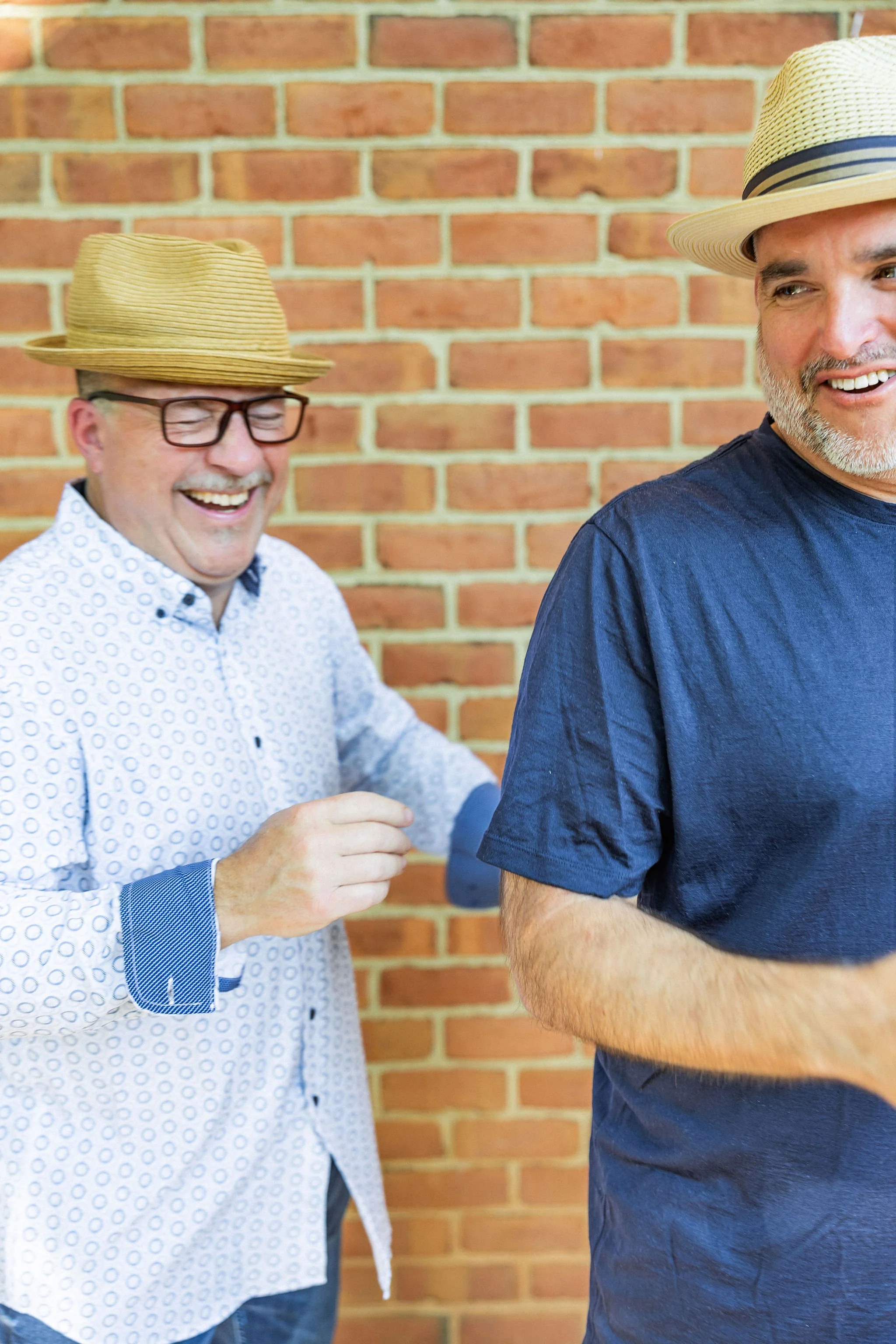 Two men smiling and laughing, both wearing straw hats, standing outdoors against a brick wall.