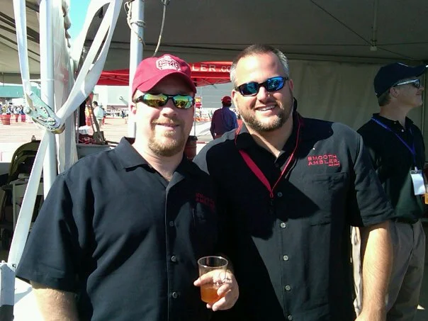Two men smiling at an outdoor event, one holding a glass of beer, both wearing black shirts with red logos, and sunglasses.