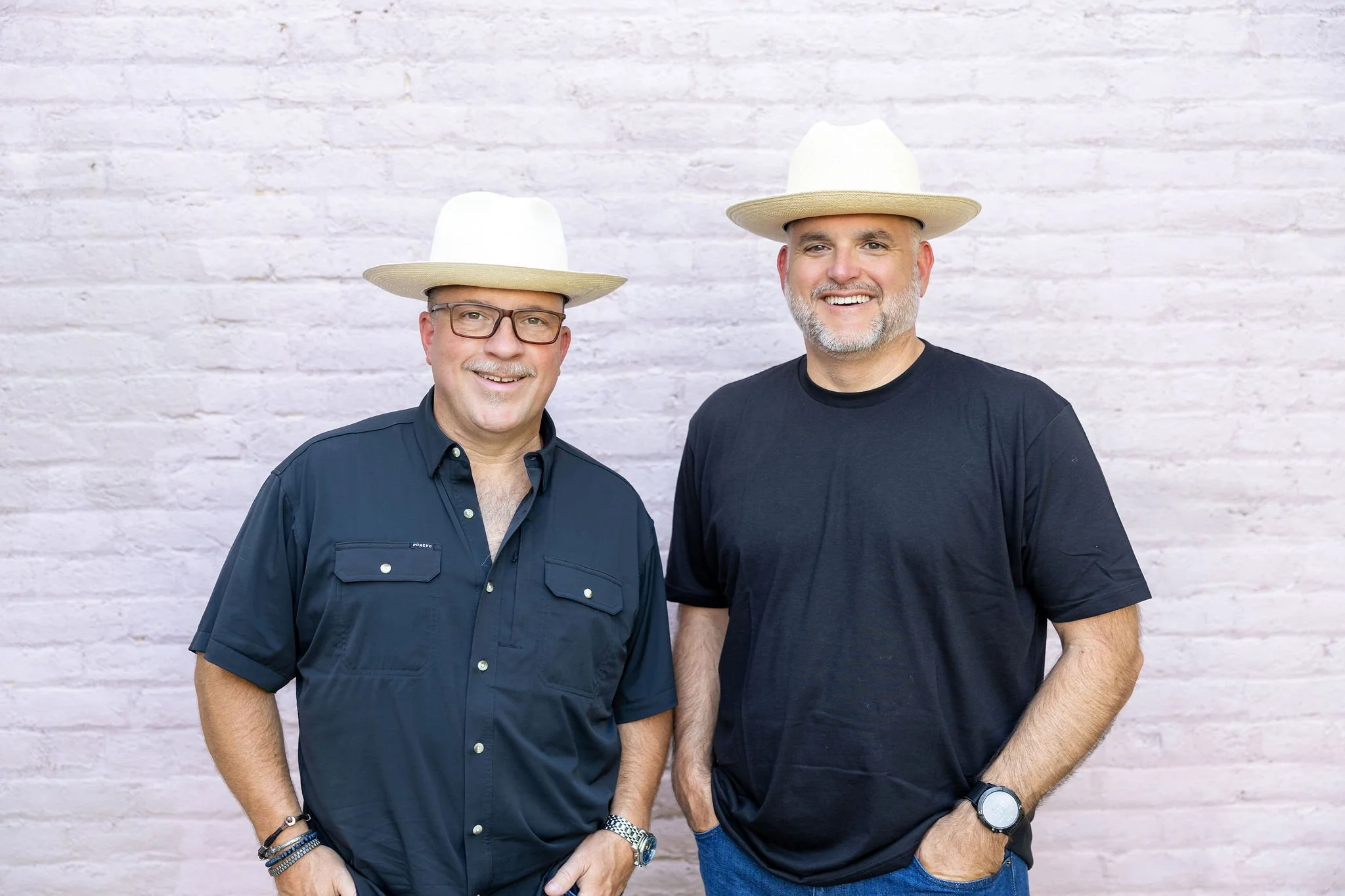 Two smiling men standing in front of a white brick wall, wearing white cowboy hats and casual clothing.
