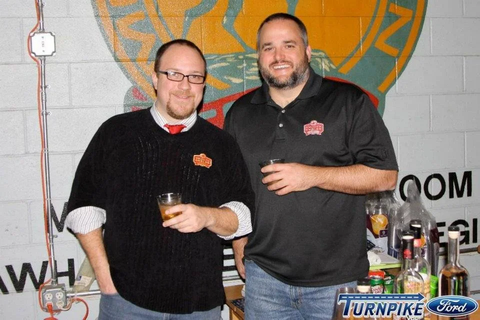 Two men standing inside a bar or brewery, holding drinks and smiling at the camera. One wears glasses, a black sweater, and a white shirt with a red tie. The other has a beard, is in a black polo shirt, and holds a glass. Background features a colorf