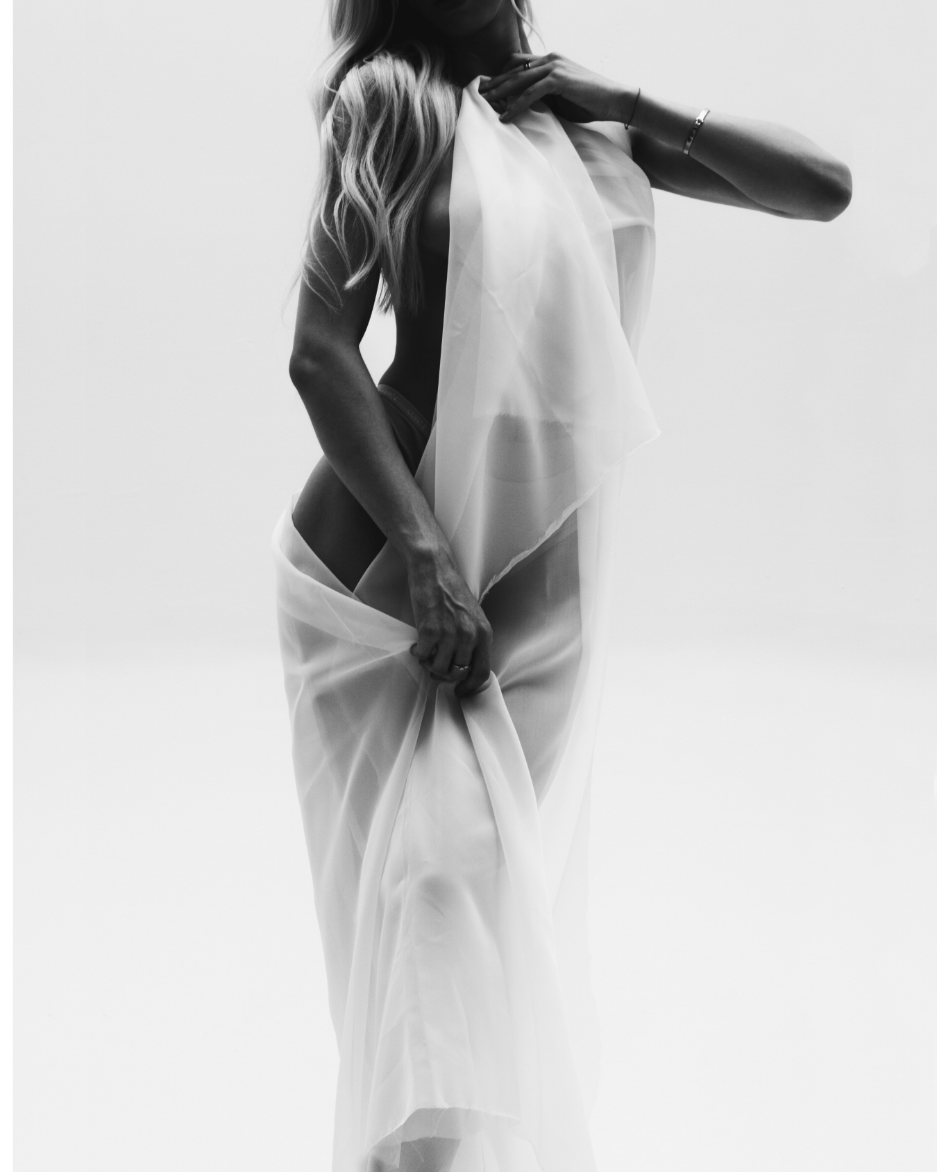 A black and white photograph of a woman in sheer fabric, partially covering her body and face, with her hair styled in loose waves.