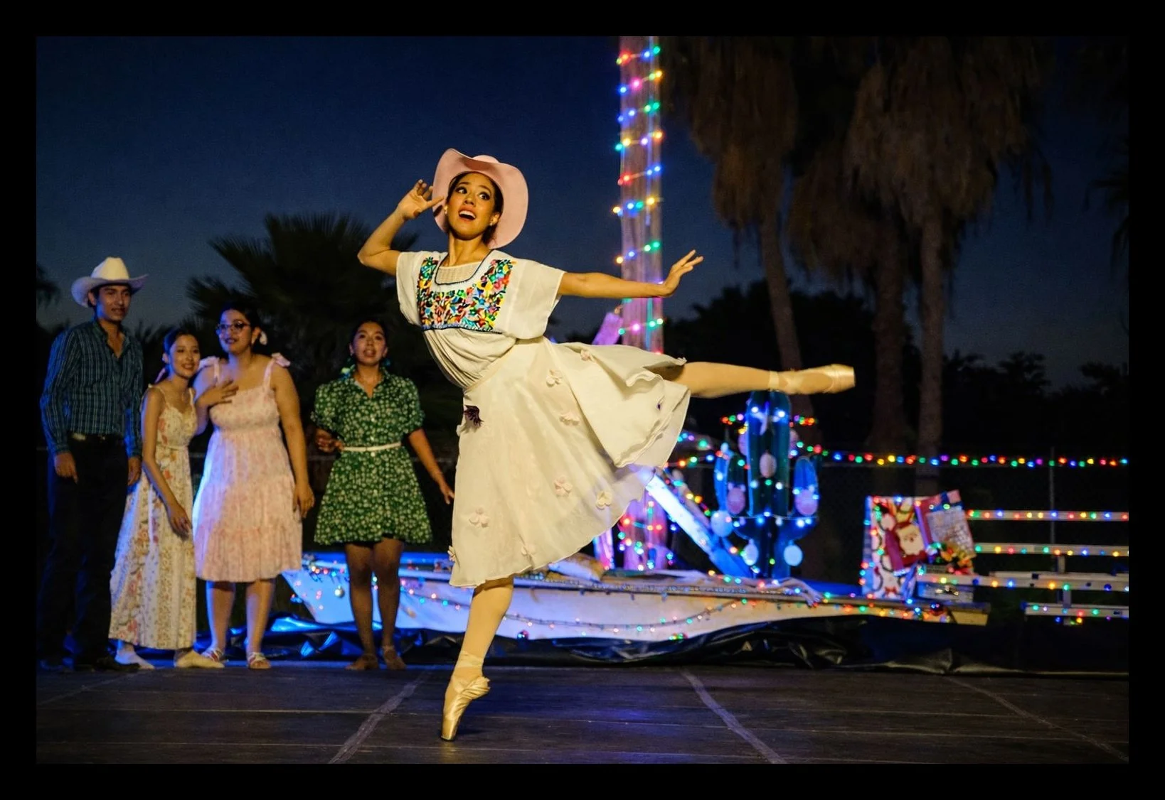 Una bailarina con vestido y sombrero rosas baila de ballet en un escenario durante la noche, mientras un grupo de personas vestidas con ropa campestre observa y la rodea, con decoraciones de luces de colores y un fondo de palmeras.