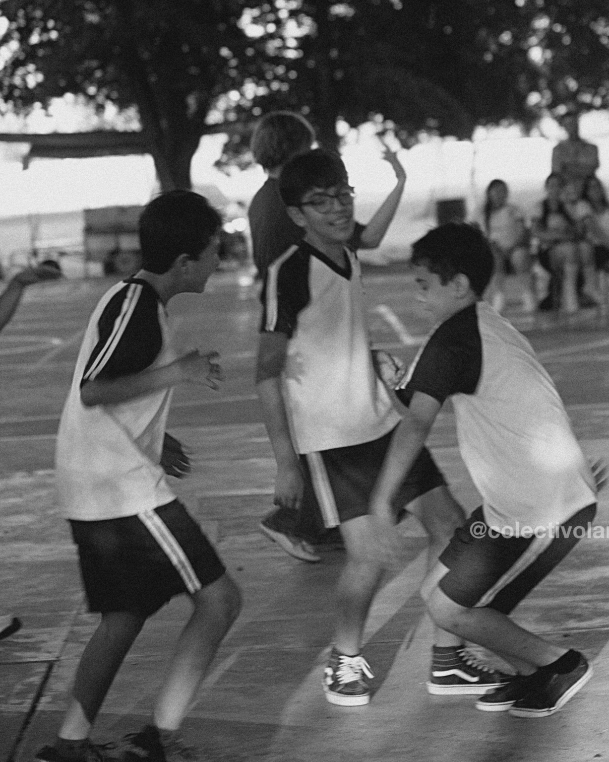 Niños jugando en una cancha al aire libre, algunos se ven riendo y disfrutando del momento, con árboles de fondo y otras personas en el área.