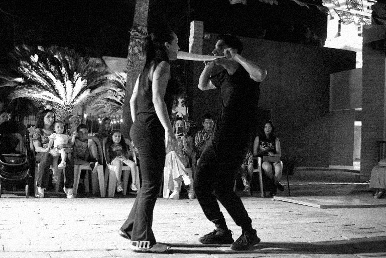 Una pareja bailando en una calle al aire libre ante un grupo de personas sentadas observando.