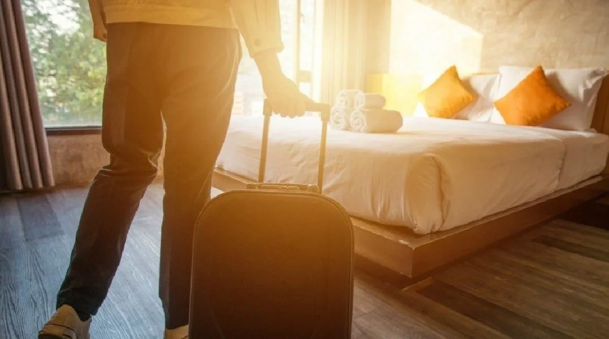 Persona con maleta en una habitación de hotel, lista para partir, con la cama hecha y toallas enrolladas en la cabecera, y luz cálida natural en la habitación.