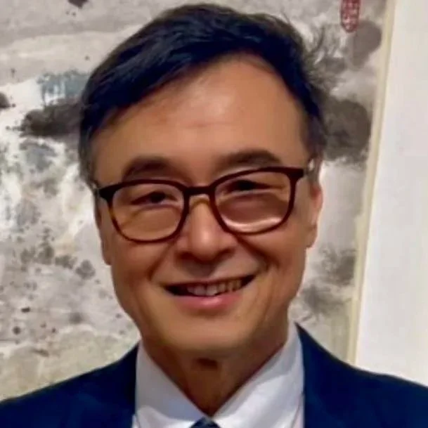 A man with short black hair, glasses, and a big smile, wearing a dark suit and white shirt, standing in front of a weathered wall.