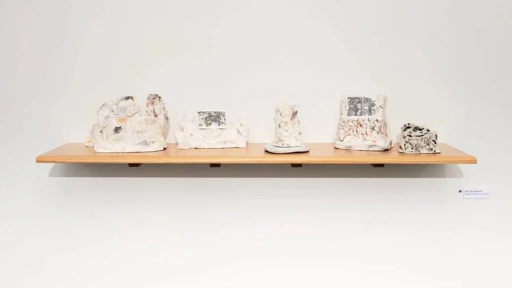 A long wooden shelf displaying five abstract art sculptures with mixed textures and images, with a small informational label on the wall to the right.