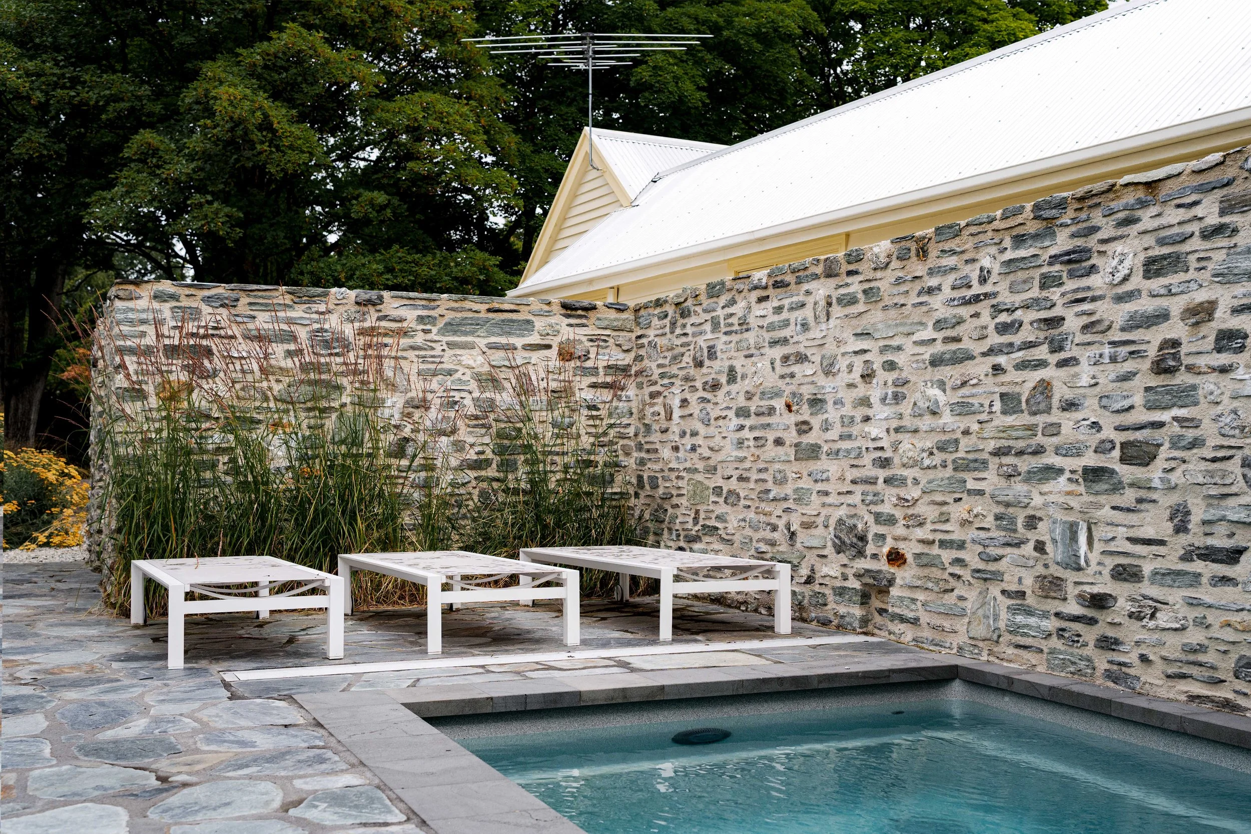 Poolside view showing yellow yarrow flowers blooming beside a plunge pool with a historic-style stone wall backdrop in Arrowtown.