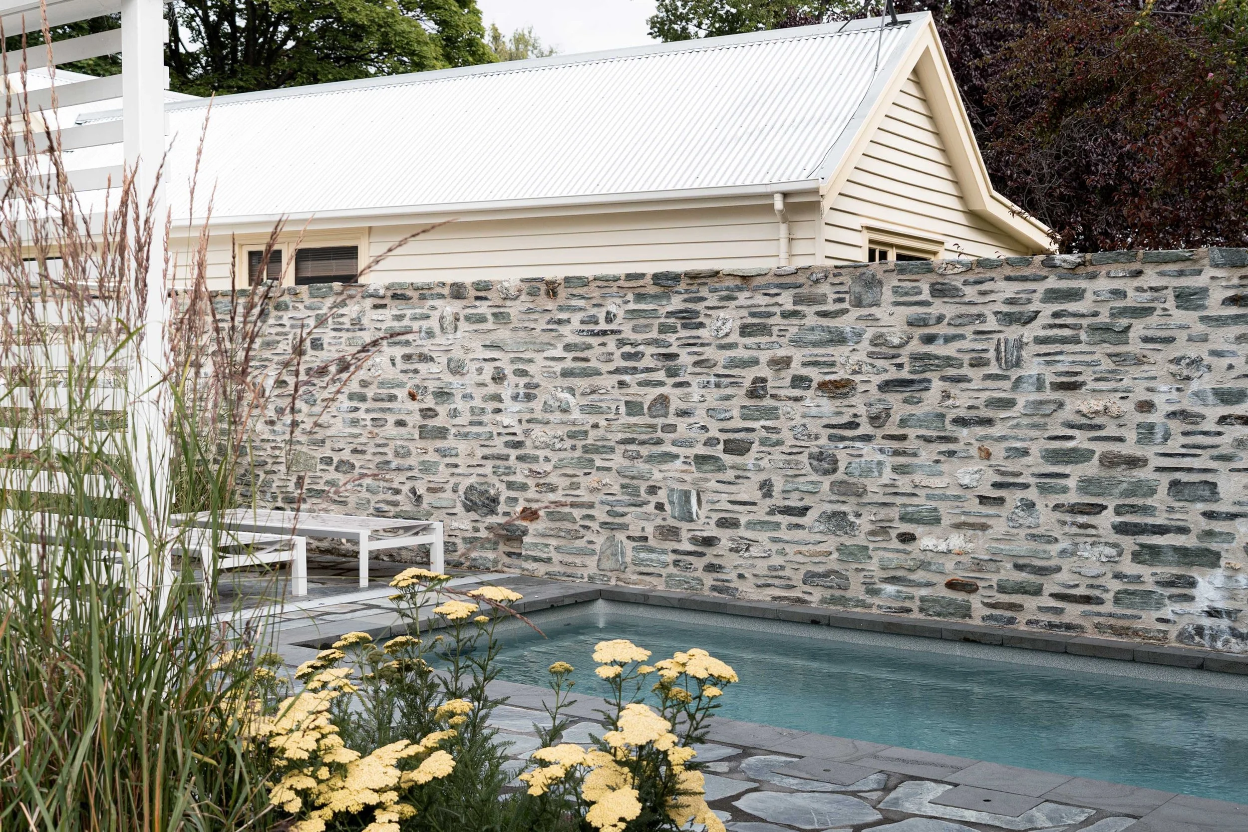 White sun loungers resting on flagstone paving behind a screen of tall feather reed grasses and a stone wall.