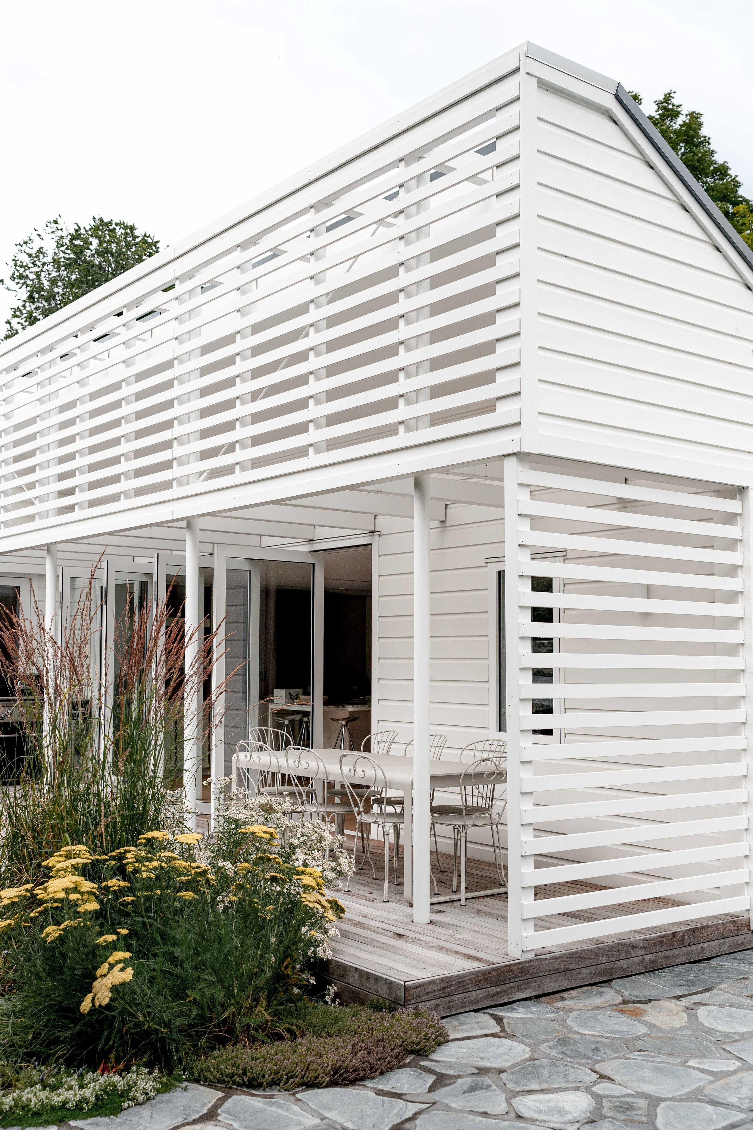 Vertical shot of a modern white slatted timber pergola attached to a white weatherboard house, sheltering an alfresco dining area.