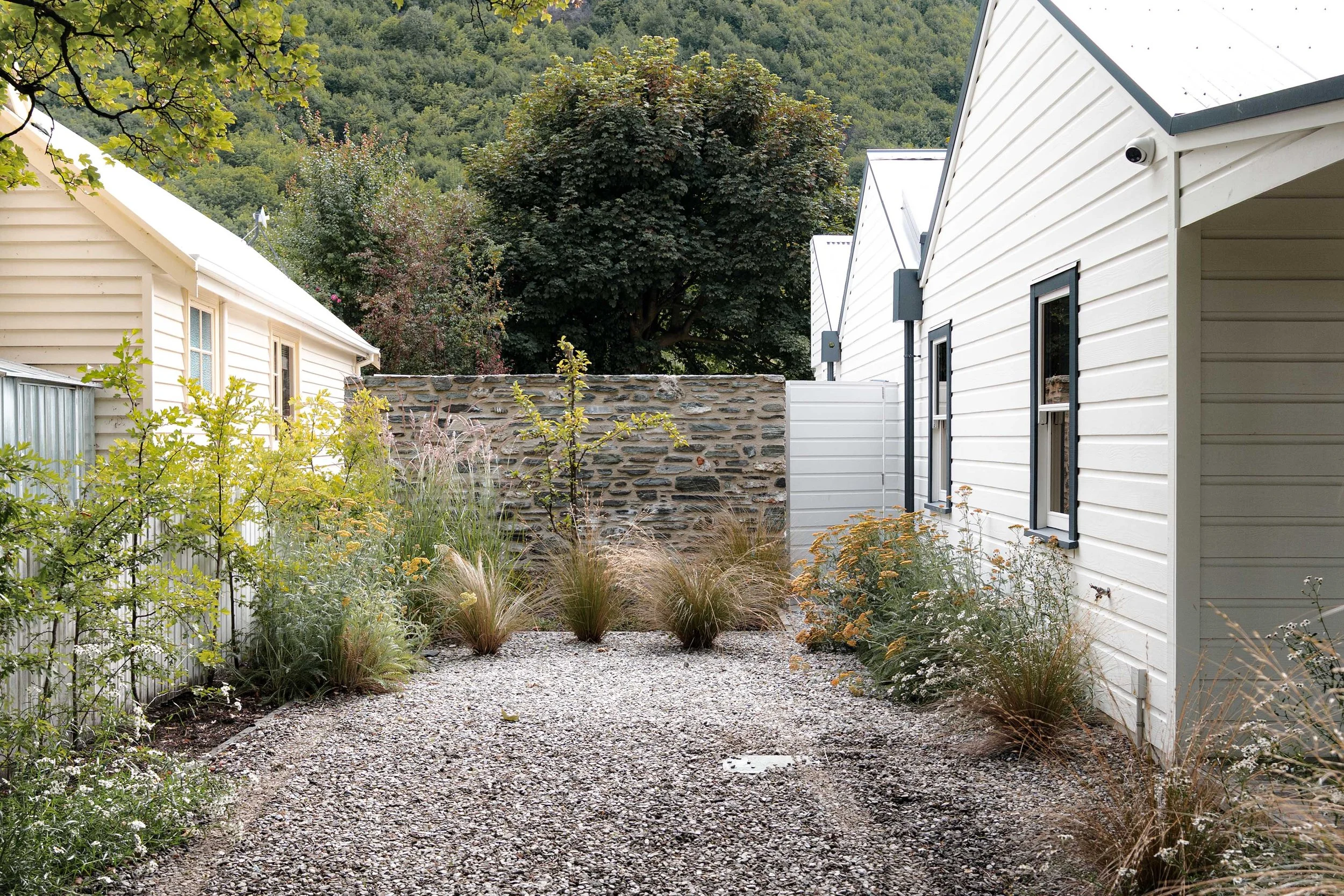 A gravel side pathway bordered by white timber siding and a traditional Central Otago schist stone wall with wild yarrow and ornamental grasses.