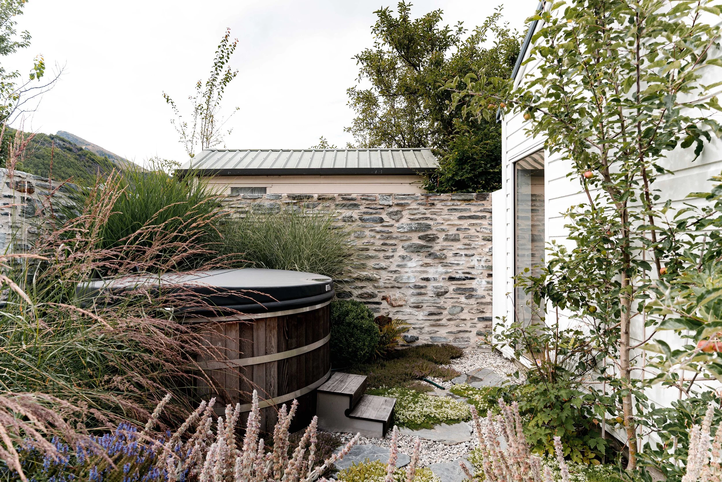 Round cedar hot tub nestled into a private courtyard garden with lavender, purple salvia, and tall grasses against a stone wall.