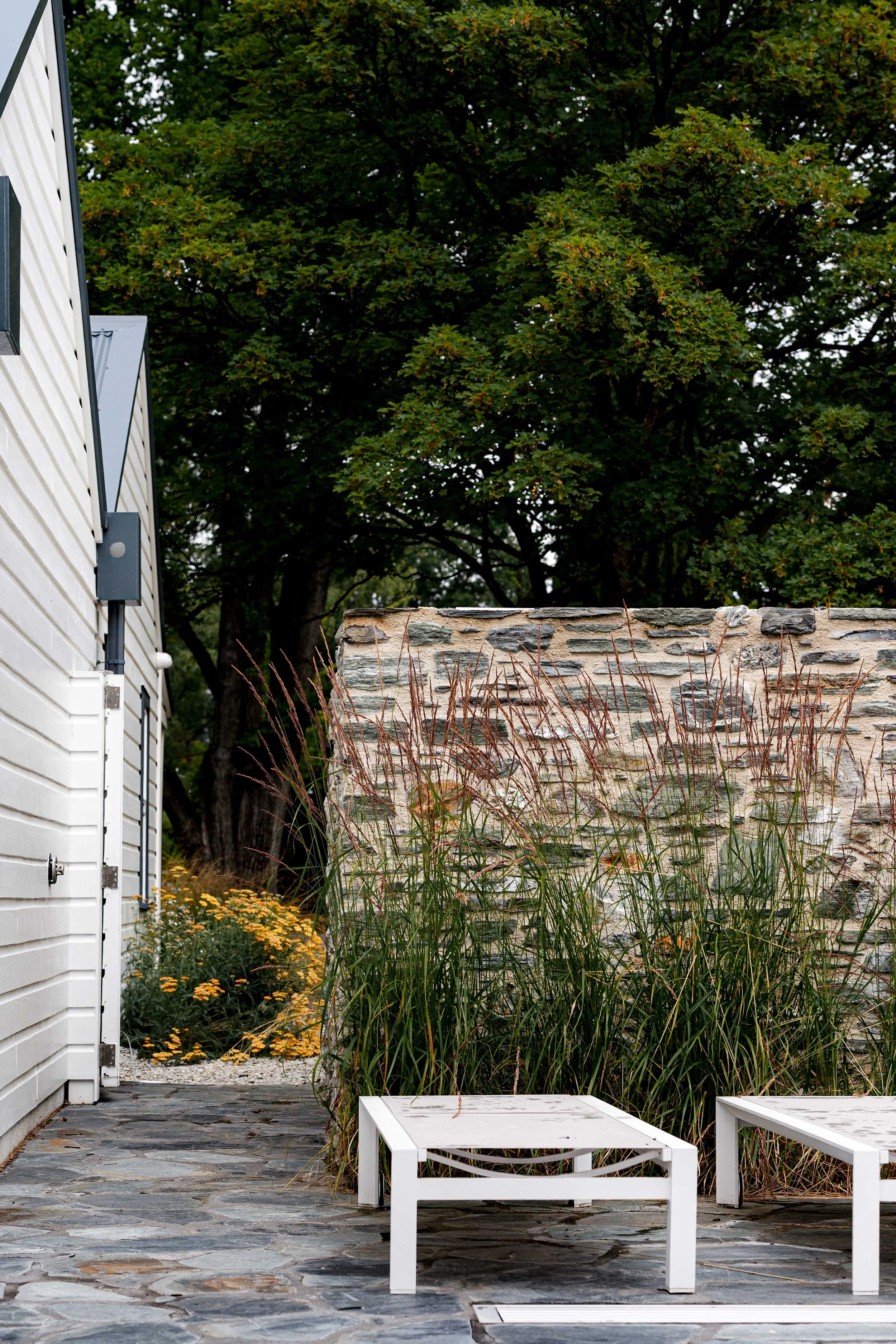 A gravel side pathway bordered by white timber siding and a traditional Central Otago schist stone wall with wild yarrow and ornamental grasses.