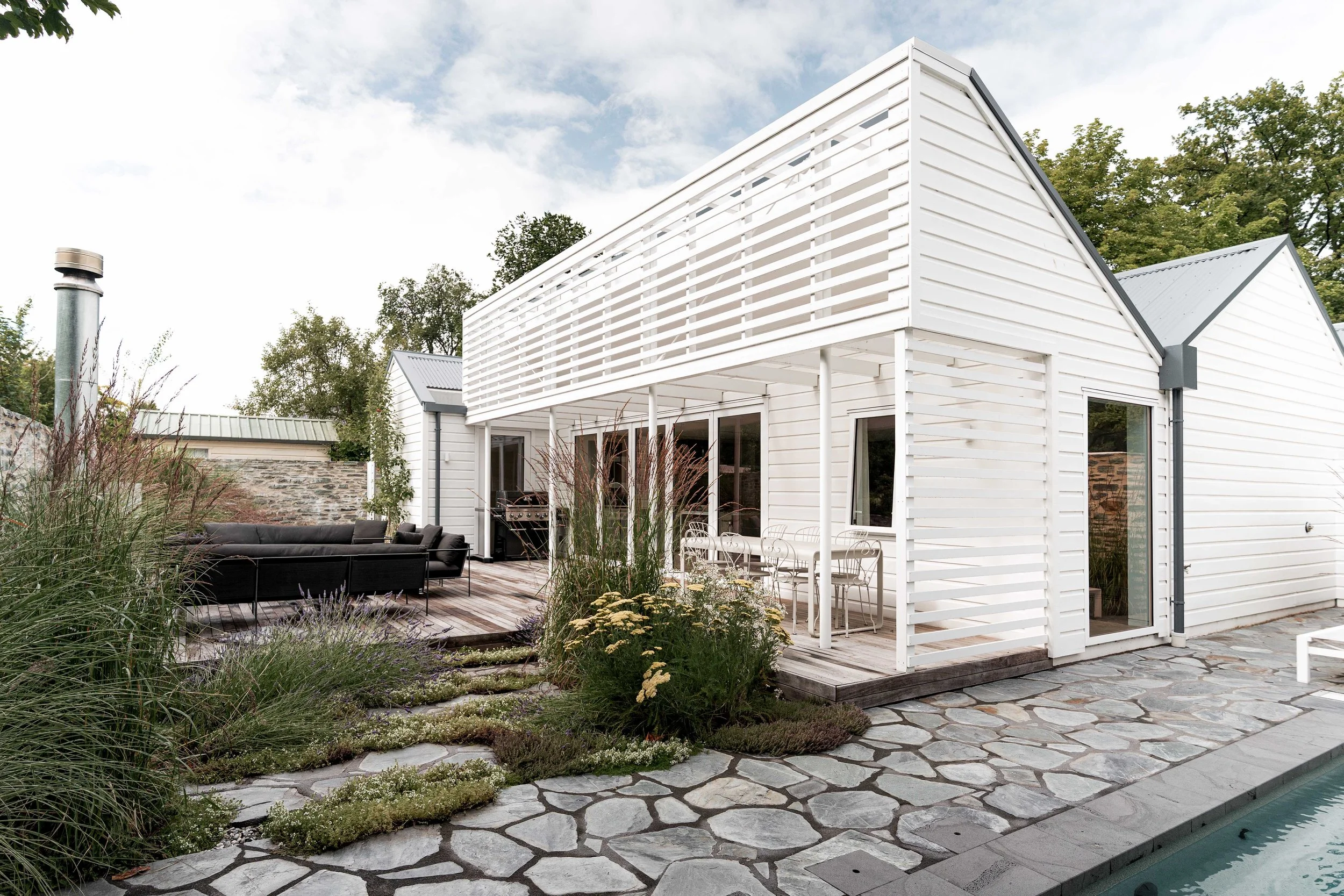 Contemporary outdoor living area featuring a white gabled building, an architectural pergola over a dining set, stone paving, and a plunge pool.