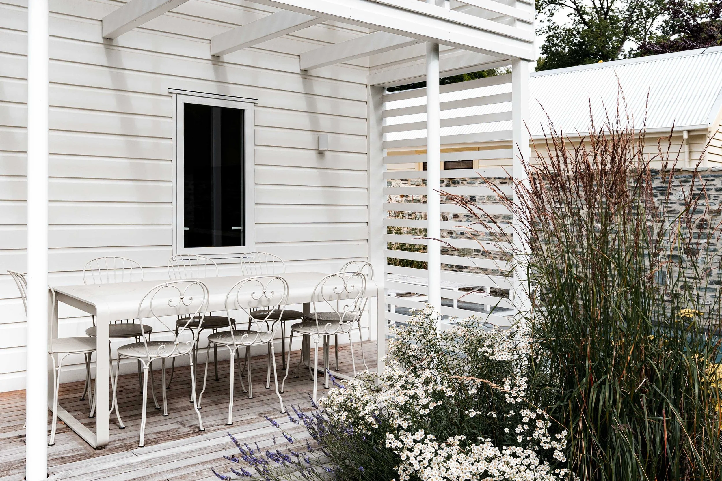 White metal outdoor dining table and classic filigree chairs positioned on a timber deck under a modern white timber pergola.