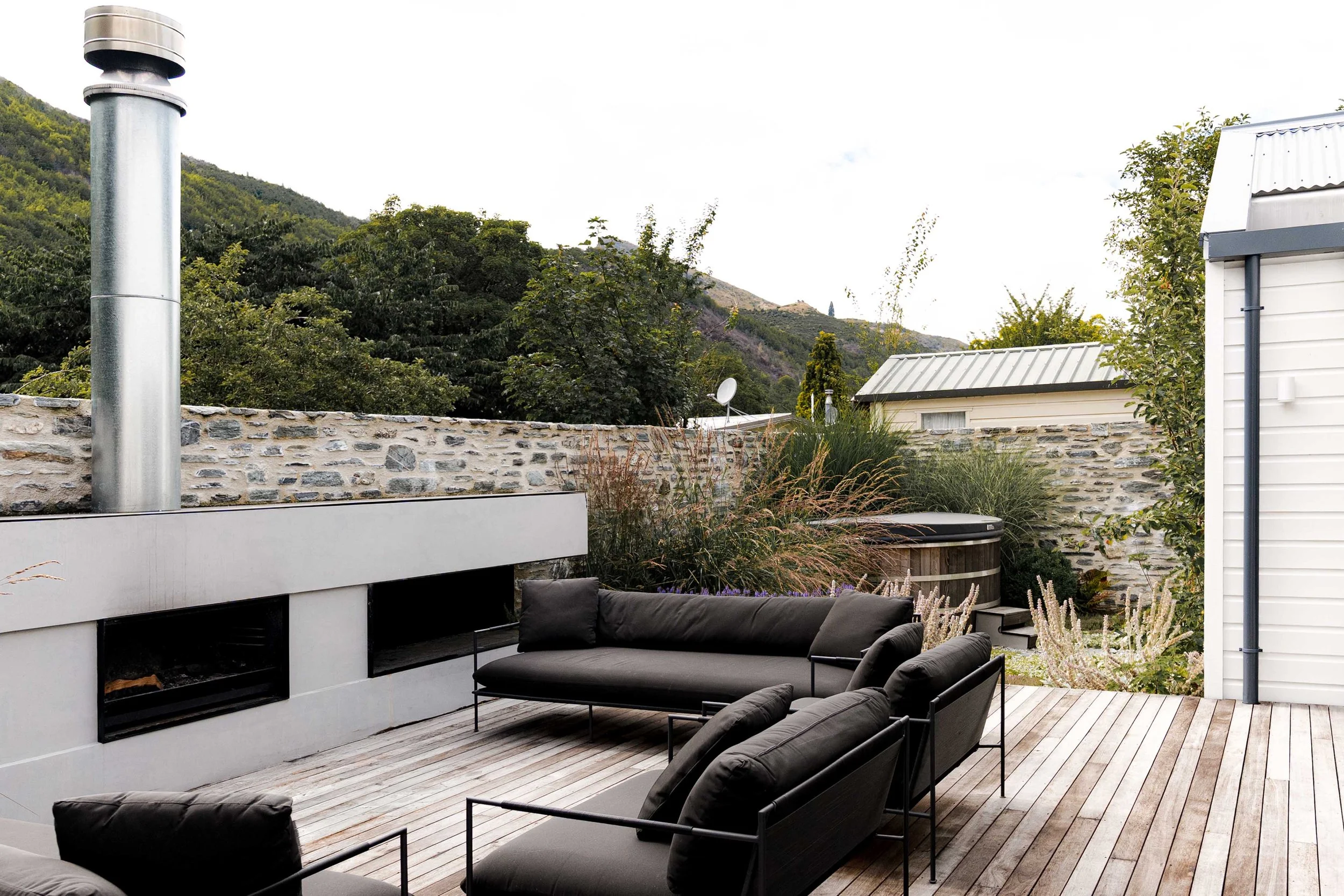 Modern black outdoor sofa layout centered around a minimalist white concrete outdoor fireplace built into a schist stone wall.