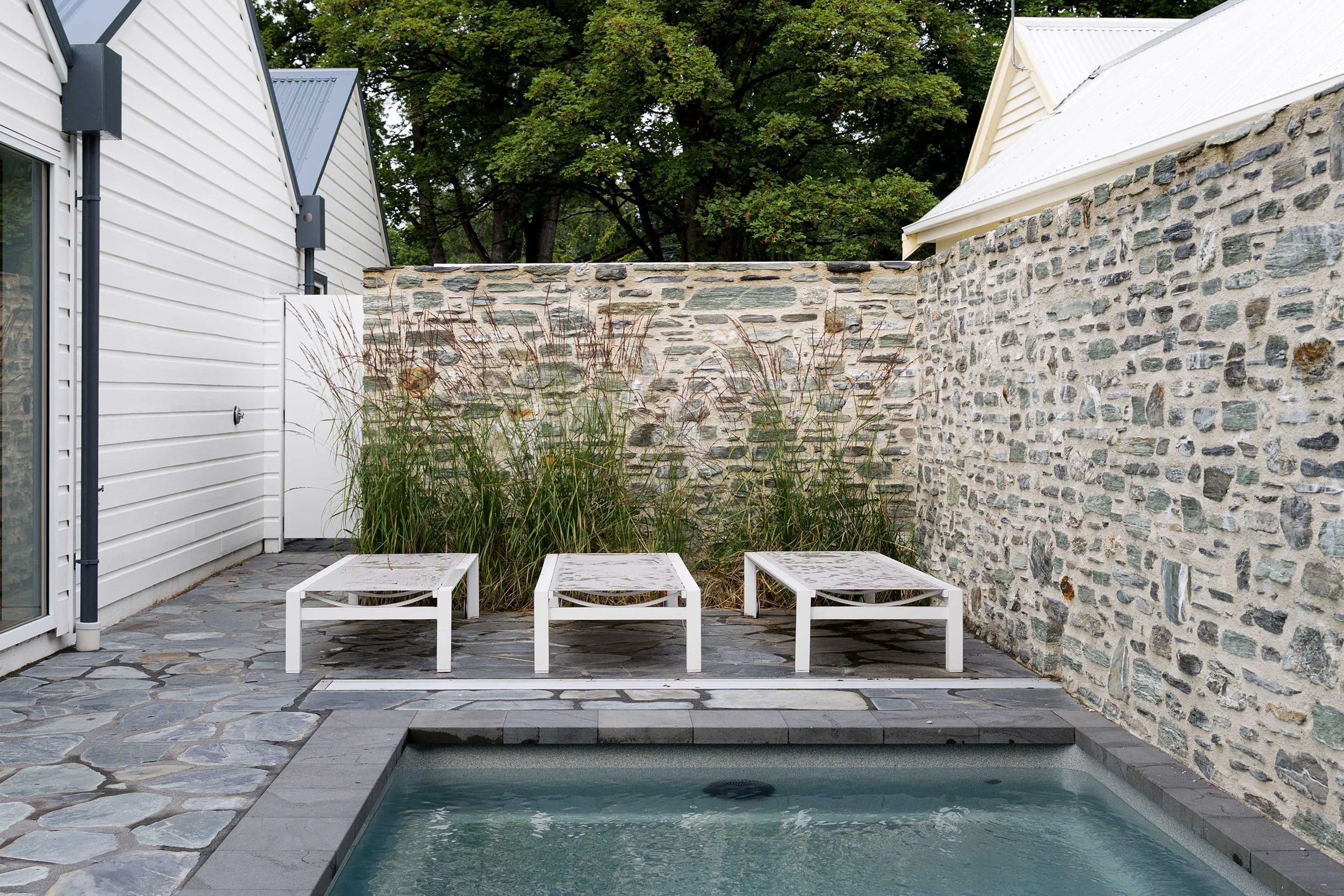 A modern rectangular plunge pool surrounded by gray stone flagstone paving, white sun loungers, and a rustic schist stone boundary wall.