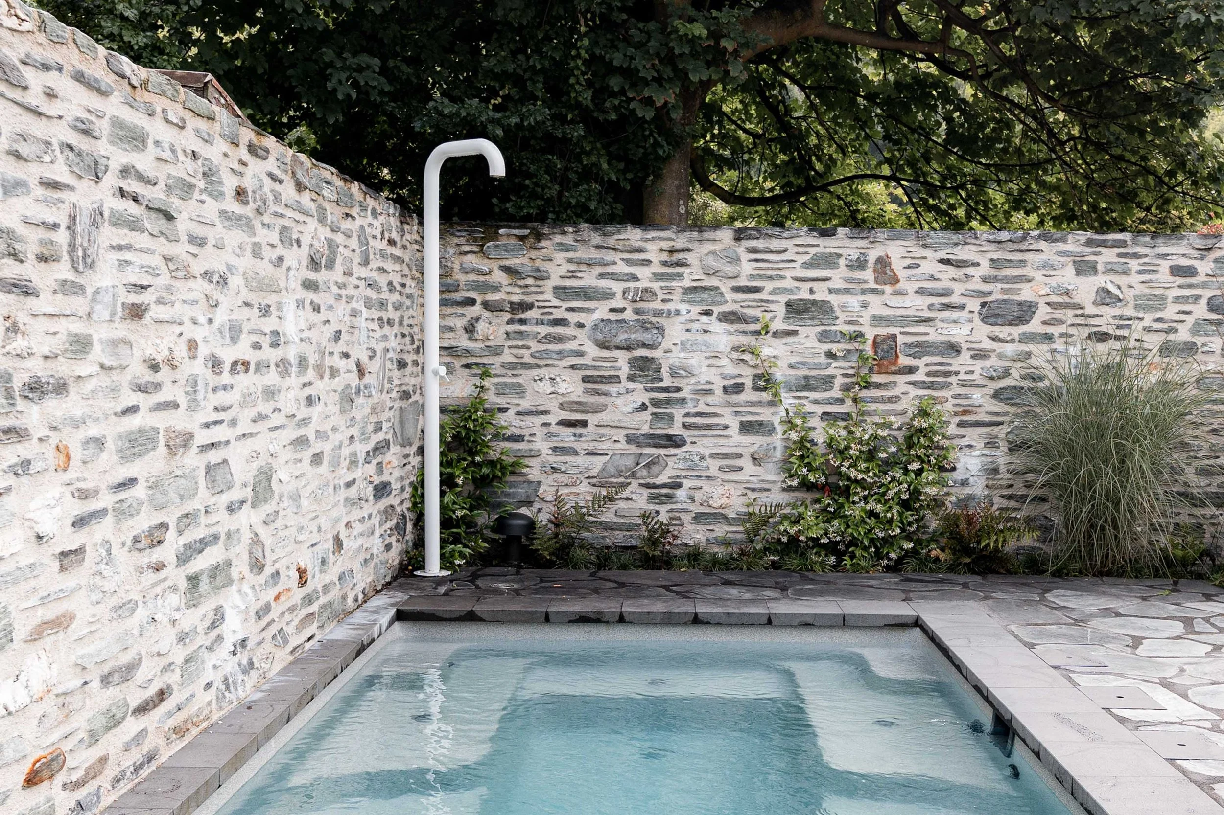 Minimalist white gooseneck outdoor shower installed on a flagstone patio next to a swimming pool and a stone wall with climbing green plants.