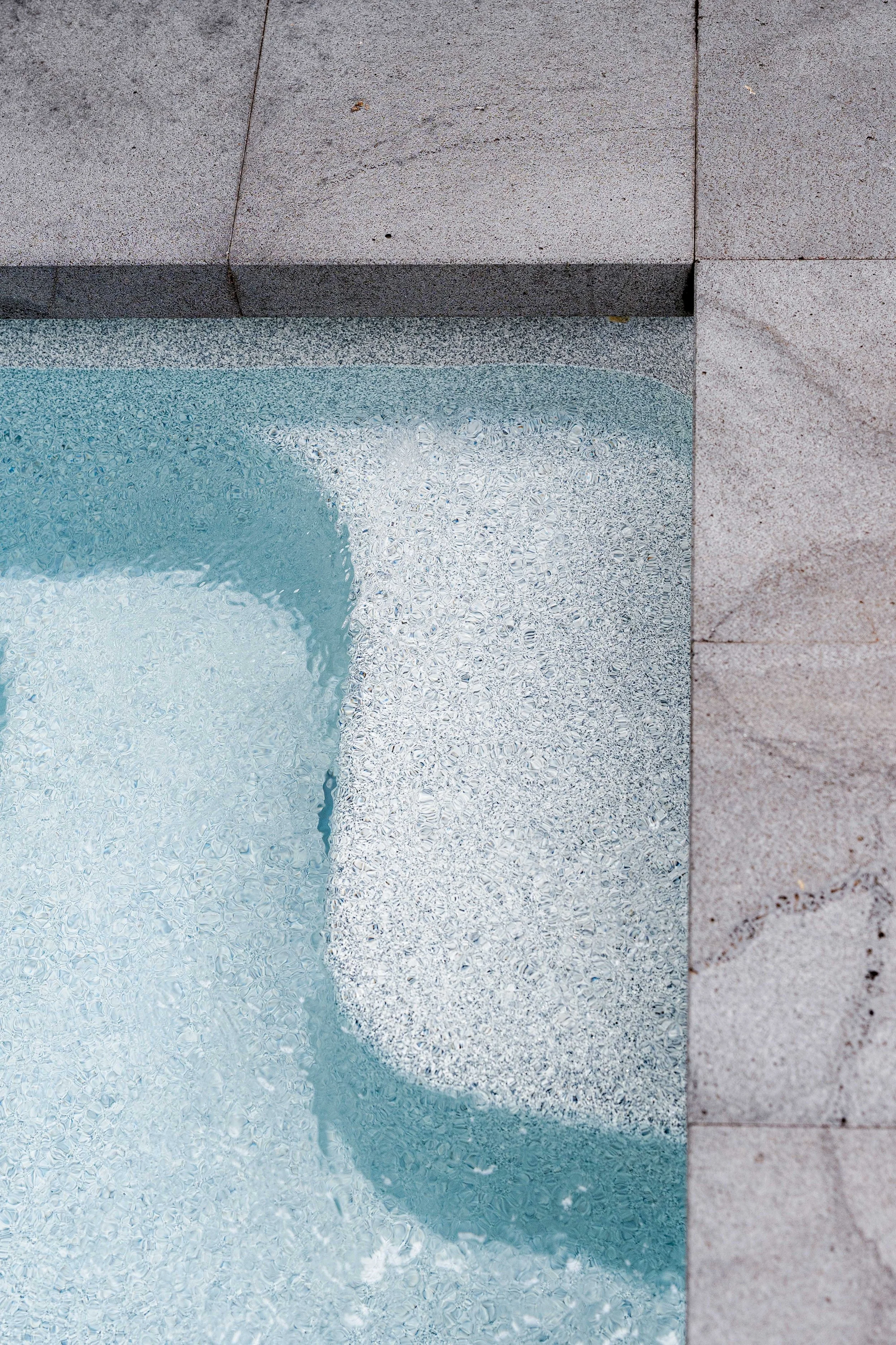 High-angle detail shot of clear blue water over a textured pool step with gray granite pool coping tiles.