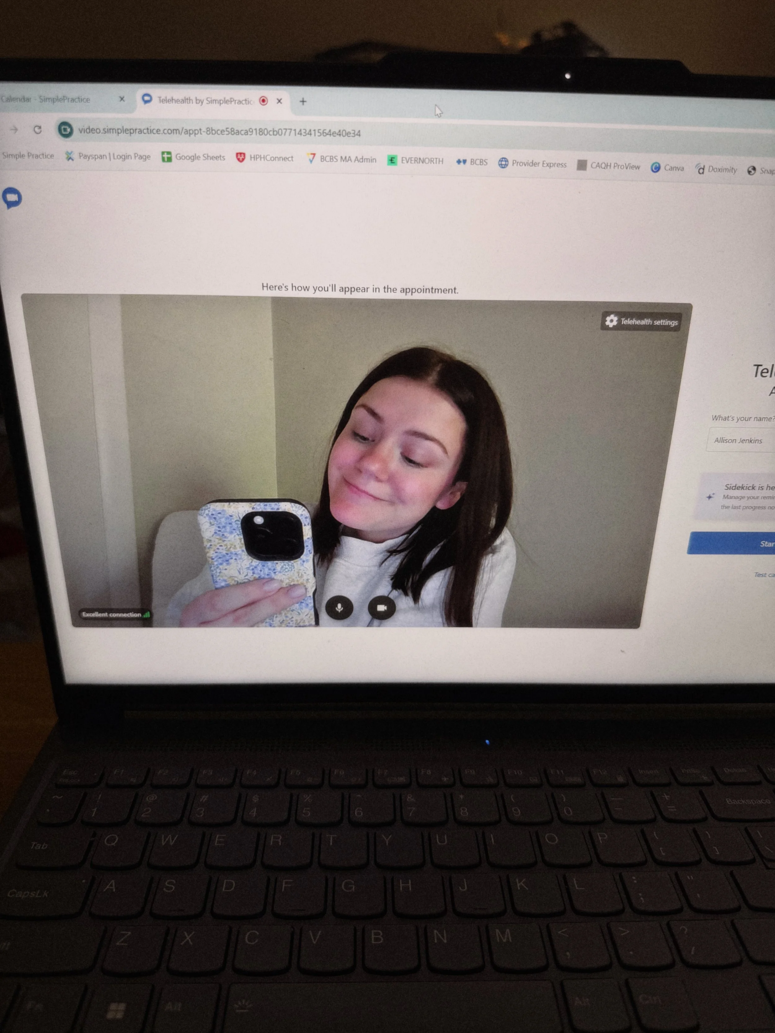 Laptop screen displaying a video call with a young woman taking a selfie, featuring dark brown hair, holding a phone, with visible keyboard in the foreground.