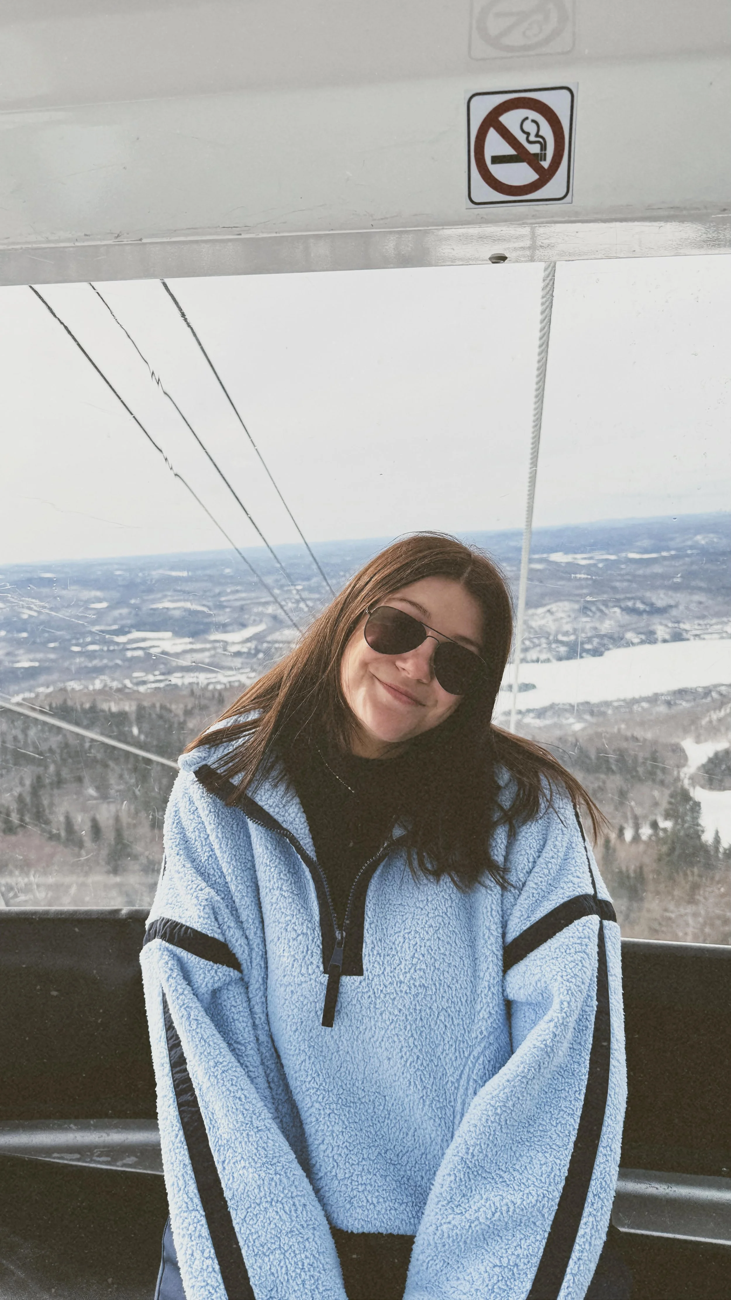 A young woman with brown hair and sunglasses smiling while standing in a cable car with a snowy landscape and lake in the background. A no smoking sign is visible above her.